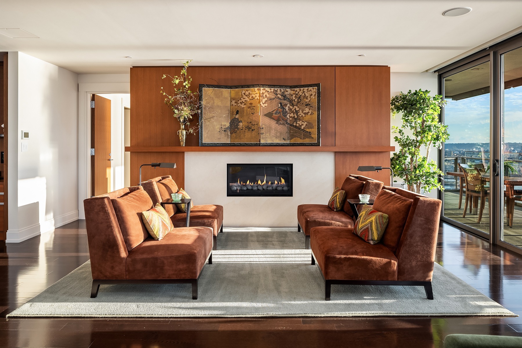 A modern living room features four brown armchairs around a coffee table facing a minimalist, wall-mounted fireplace. The backdrop is a wooden panel with an artistic painting. Large windows on the right offer a view and bring in natural light.