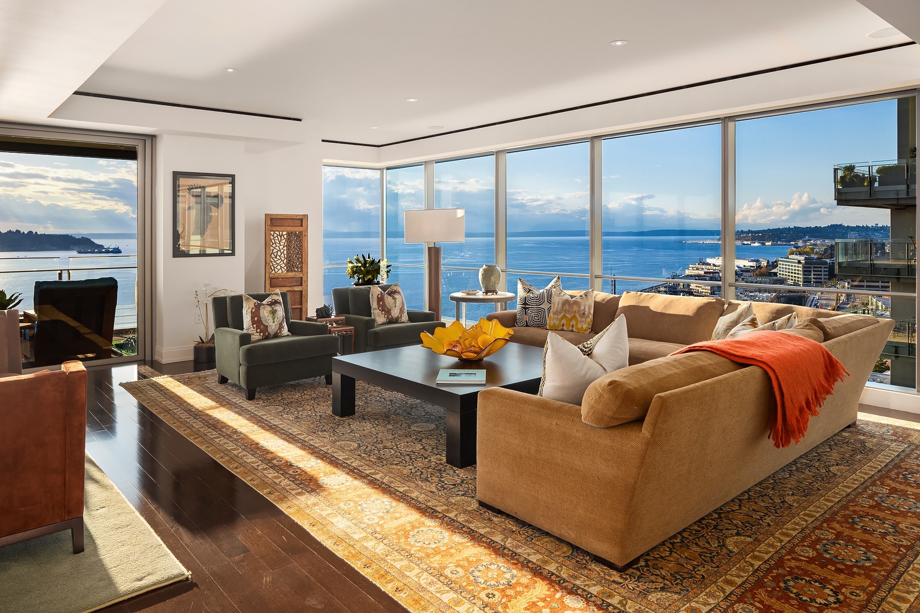 A modern living room with large windows offering a view of a waterfront and cityscape. It features a tan sectional sofa, green armchair, and a patterned rug. A wooden coffee table with decorative items sits in the center. A mix of contemporary and classic decor fills the room.