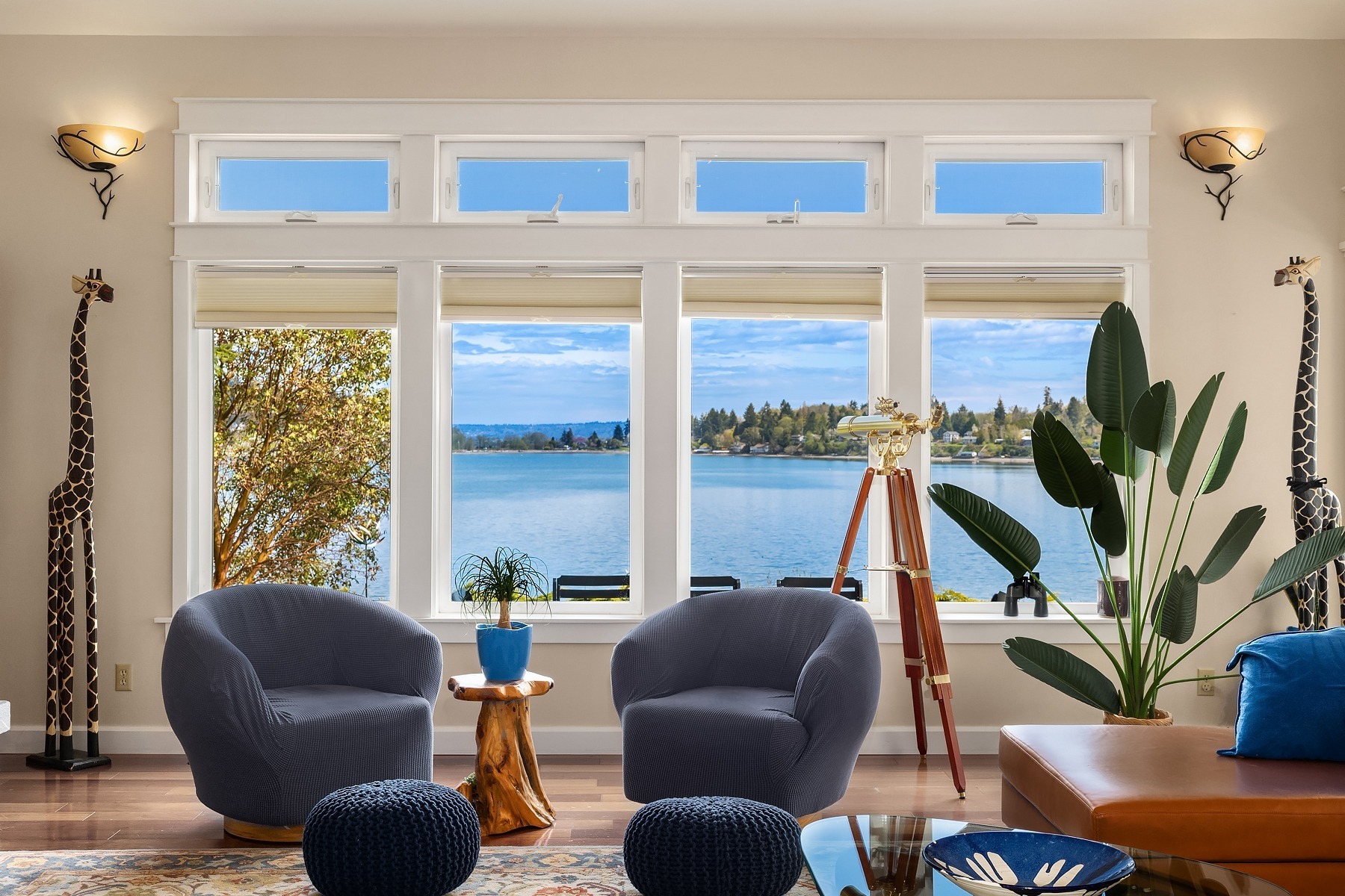 A living room with large windows overlooking a serene lake. The room features two gray chairs, a wooden side table with a blue vase, a telescope, a potted plant, giraffe statues, and blue cushions. The decor is modern, with light walls and wooden flooring.