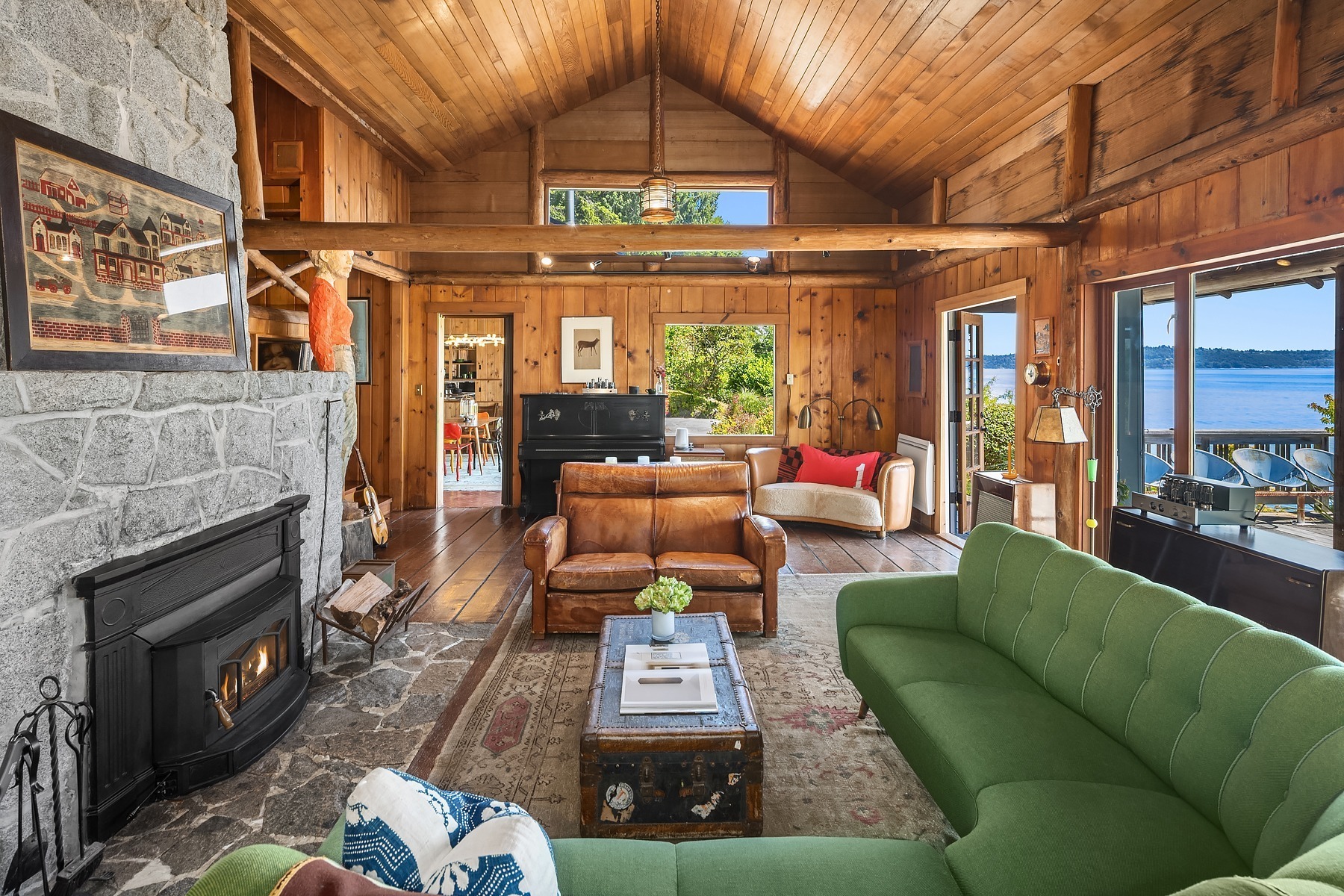 A cozy rustic living room features wooden walls and ceilings, a stone fireplace, and various comfortable seating options including green and brown sofas. Large windows and a door provide a stunning view of a lake, letting in ample natural light.