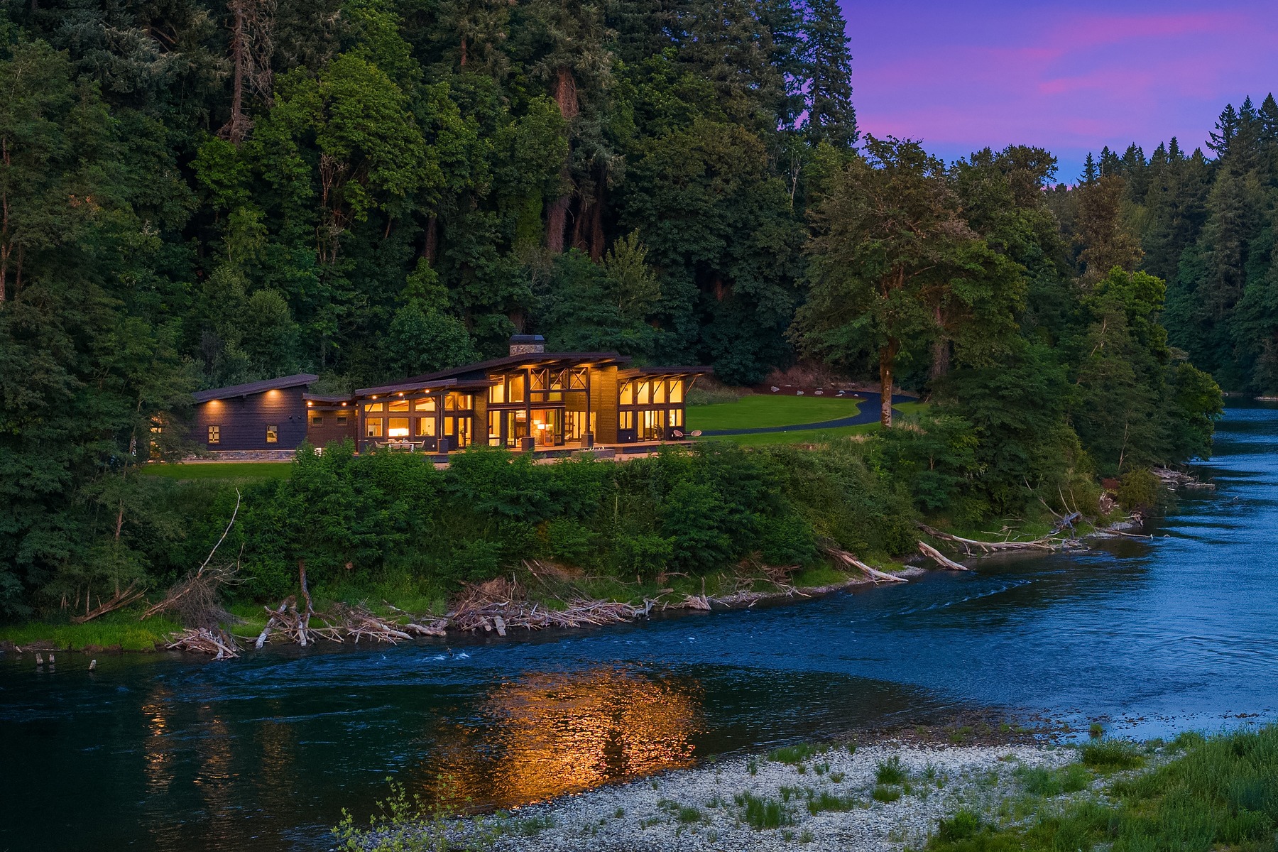 A modern house with large windows illuminated warmly inside, set amidst a lush forest and beside a serene river. The evening sky is a mix of purples and blues, reflecting on the water. Logs and branches are scattered along the riverbank, enhancing the natural setting.