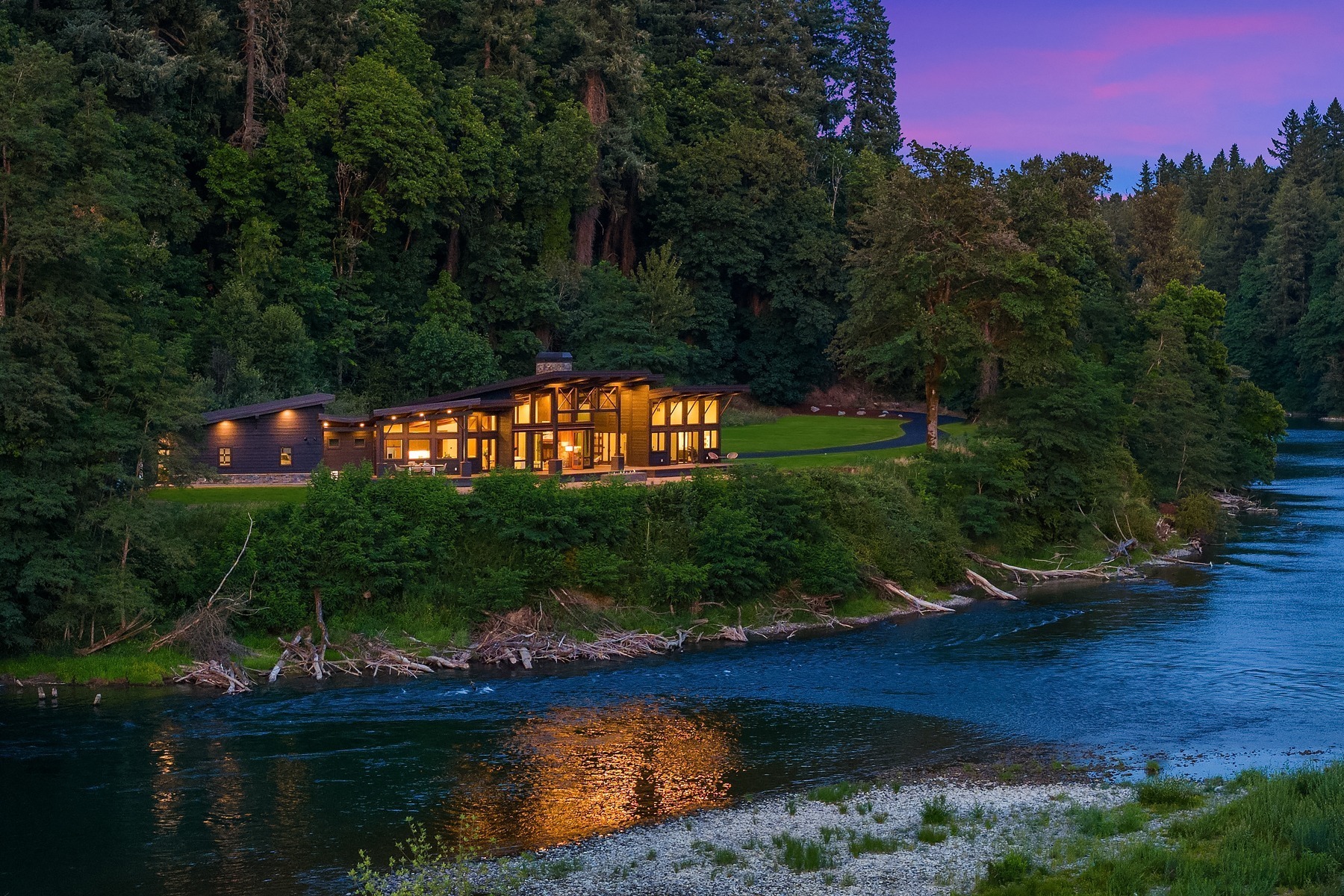 A modern, well-lit house with large windows stands on a lush green hill next to a calm river. Tall trees surround the house and line the riverbanks. The sky is purple, suggesting dusk or dawn, with the house’s warm lights reflecting in the water.