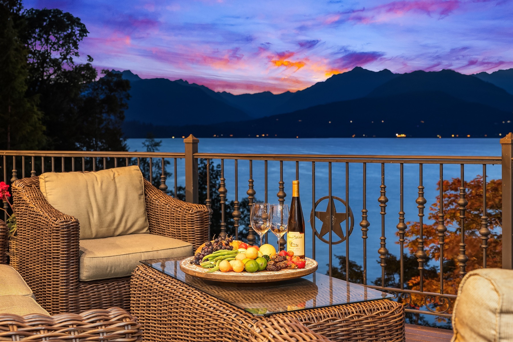 A cozy outdoor seating area with rattan furniture overlooking a serene lake and mountain range at sunset. A glass coffee table holds a wine bottle, two glasses, and a platter of assorted fruits. The sky is painted with vibrant hues of orange, pink, and purple.