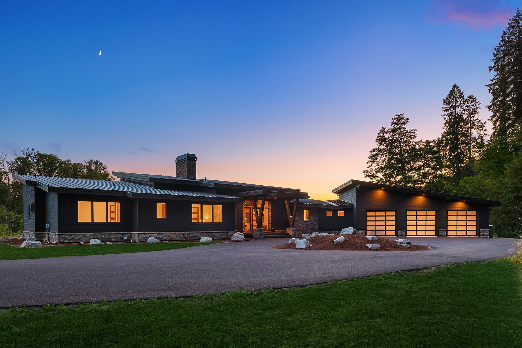 Modern single-story house with large windows illuminated from within at sunset. The home features a sleek, dark exterior and adjacent garage with multiple glass-paneled doors. A smooth driveway curves in front, surrounded by a lush green lawn and trees. A crescent moon is visible in the sky.