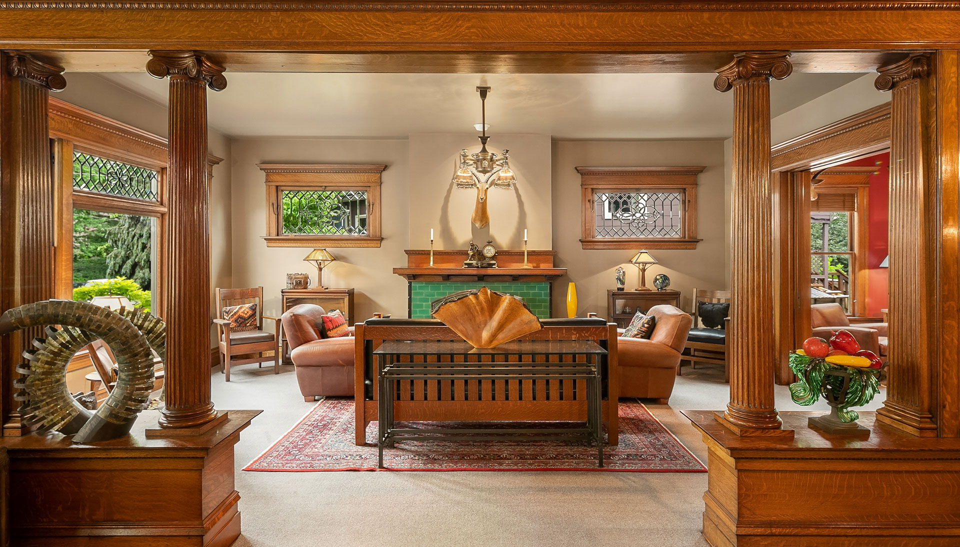 A cozy living room features wooden columns and trim, with a central wooden bench and comfortable armchairs arranged around a green-tiled fireplace. The room is decorated with intricate stained glass windows, art pieces, and warm lighting from table lamps.