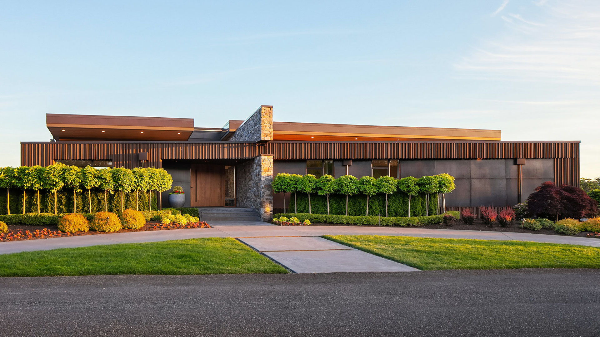 A modern, single-story house with a flat roof, nestled behind a well-manicured garden. The exterior is a mix of dark wood and stone, featuring large windows. The front yard has a variety of shrubs and a neatly trimmed lawn, with a paved walkway leading to the entrance.