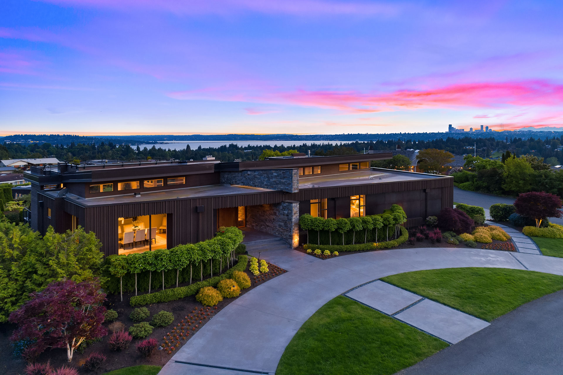 A modern, brown-stoned house with large windows, surrounded by lush green landscaping and trees. A curved driveway leads up to the entrance. The background features a vibrant sunset sky with pink and purple hues, a distant body of water, and a city skyline.