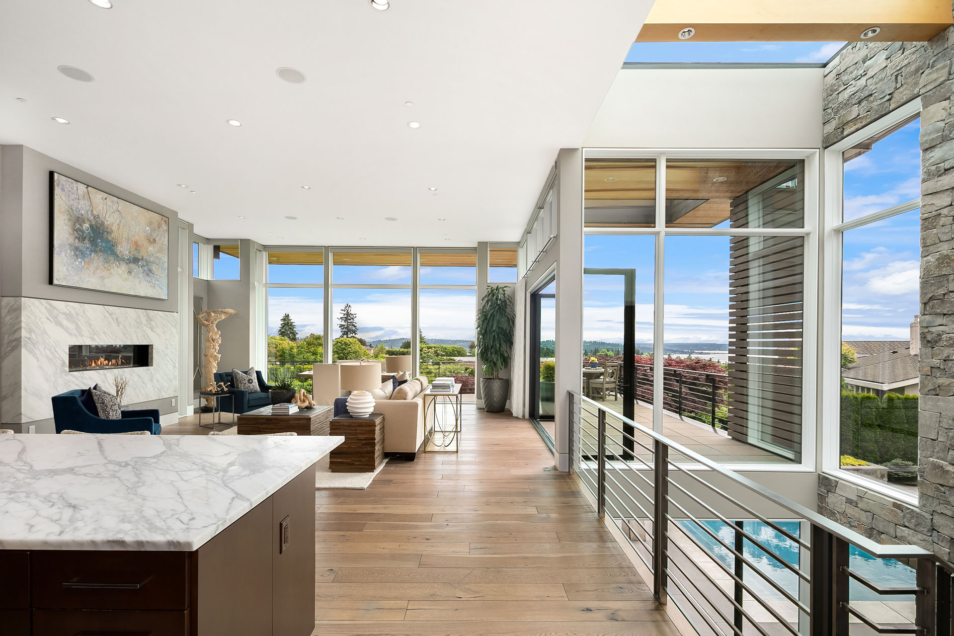 A modern, open-concept living room with floor-to-ceiling windows showcasing a scenic view. The room features hardwood floors, a marble kitchen island, contemporary furniture, a stone accent wall, and a fireplace. The space is filled with natural light.