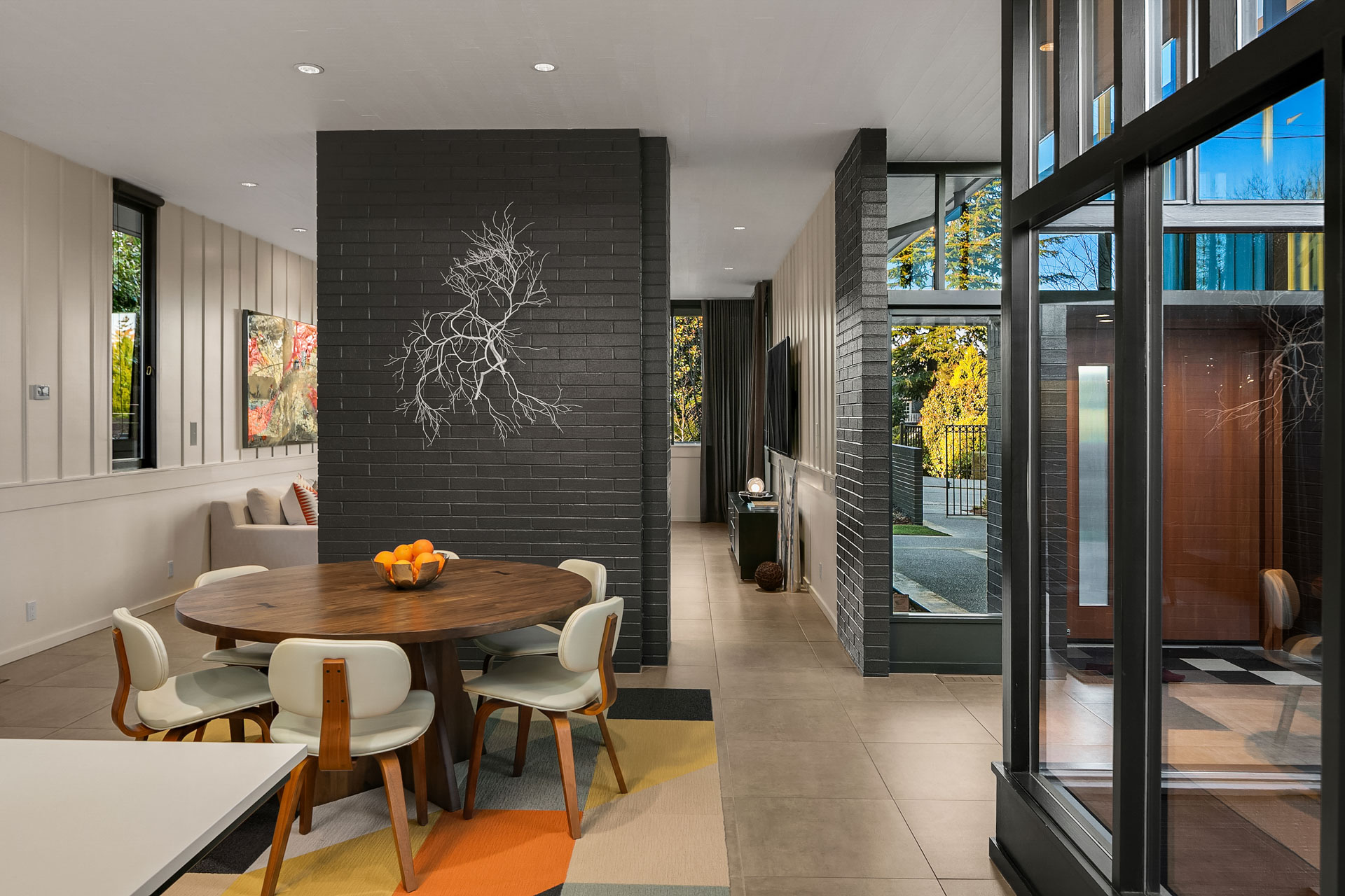 A modern dining area featuring a round wooden table with four white chairs. A minimalist centerpiece with oranges is on the table. The room has large windows letting in natural light, and a black brick accent wall with intricate wall art.