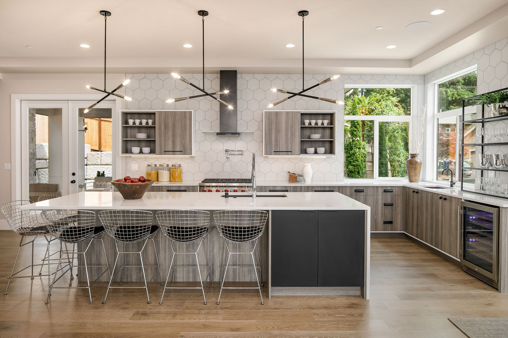 Modern kitchen with a large island featuring four wireframe barstools. The space is well-lit by contemporary pendant lights and large windows. The kitchen has gray cabinets, white hexagonal-tile backsplash, stainless steel appliances, and a built-in wine fridge.