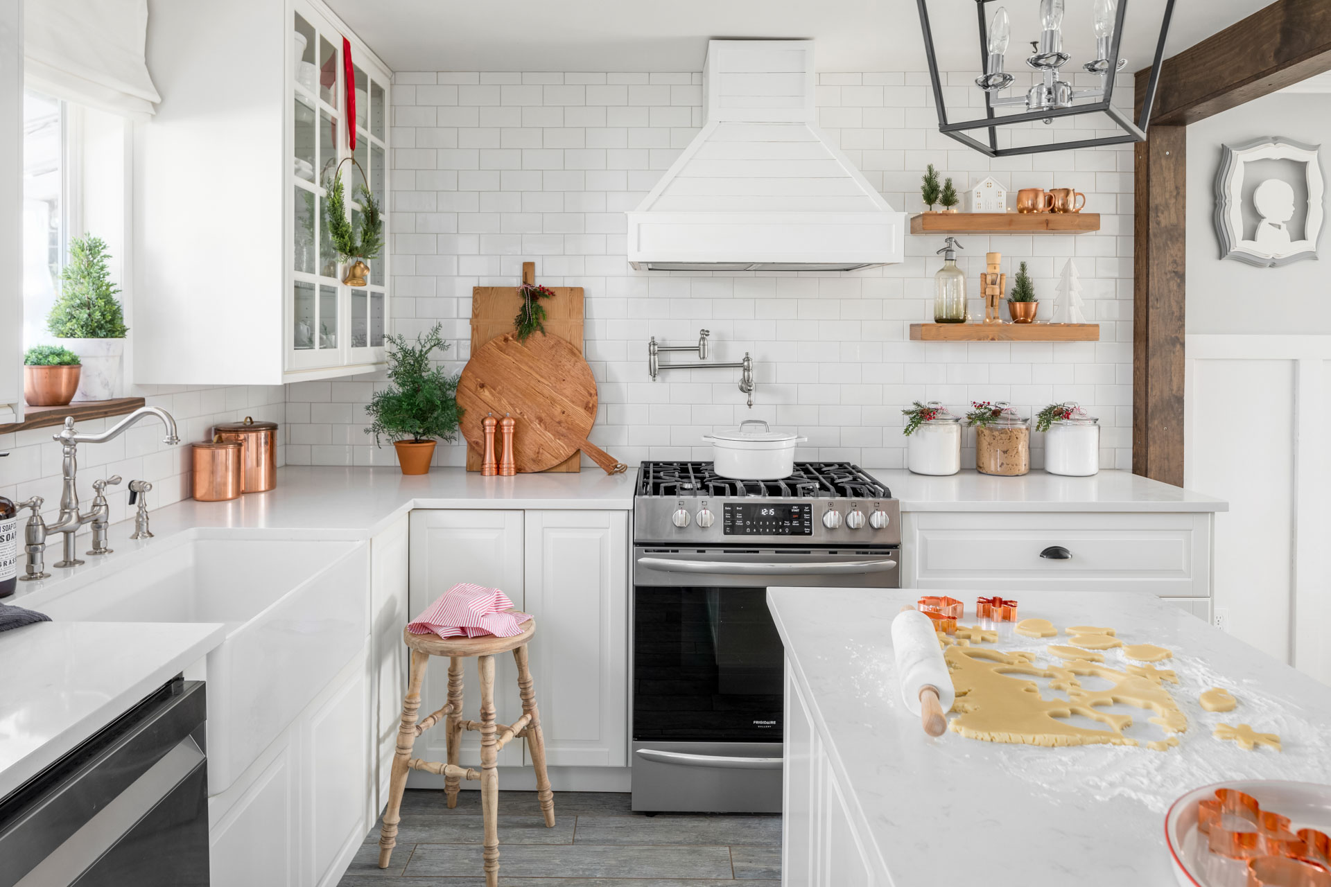 A bright, modern kitchen with white cabinetry, a farmhouse sink, and white subway tile backsplash. A stove with a white range hood is centered, with a wooden cutting board and pots on countertops. An island features a rolling pin and cookie dough being prepared.