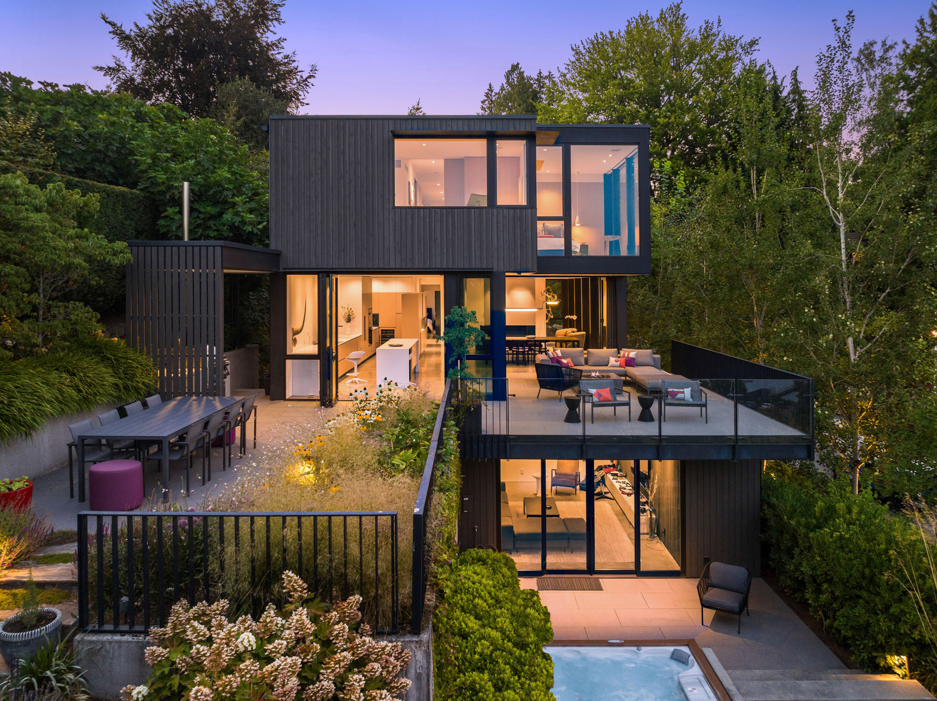 Modern two-story house with large windows and a minimalist design. The outdoor area features a spacious patio with seating, a dining table, and a hot tub. Lush greenery surrounds the property, and the interior is visible through the expansive glass panels.