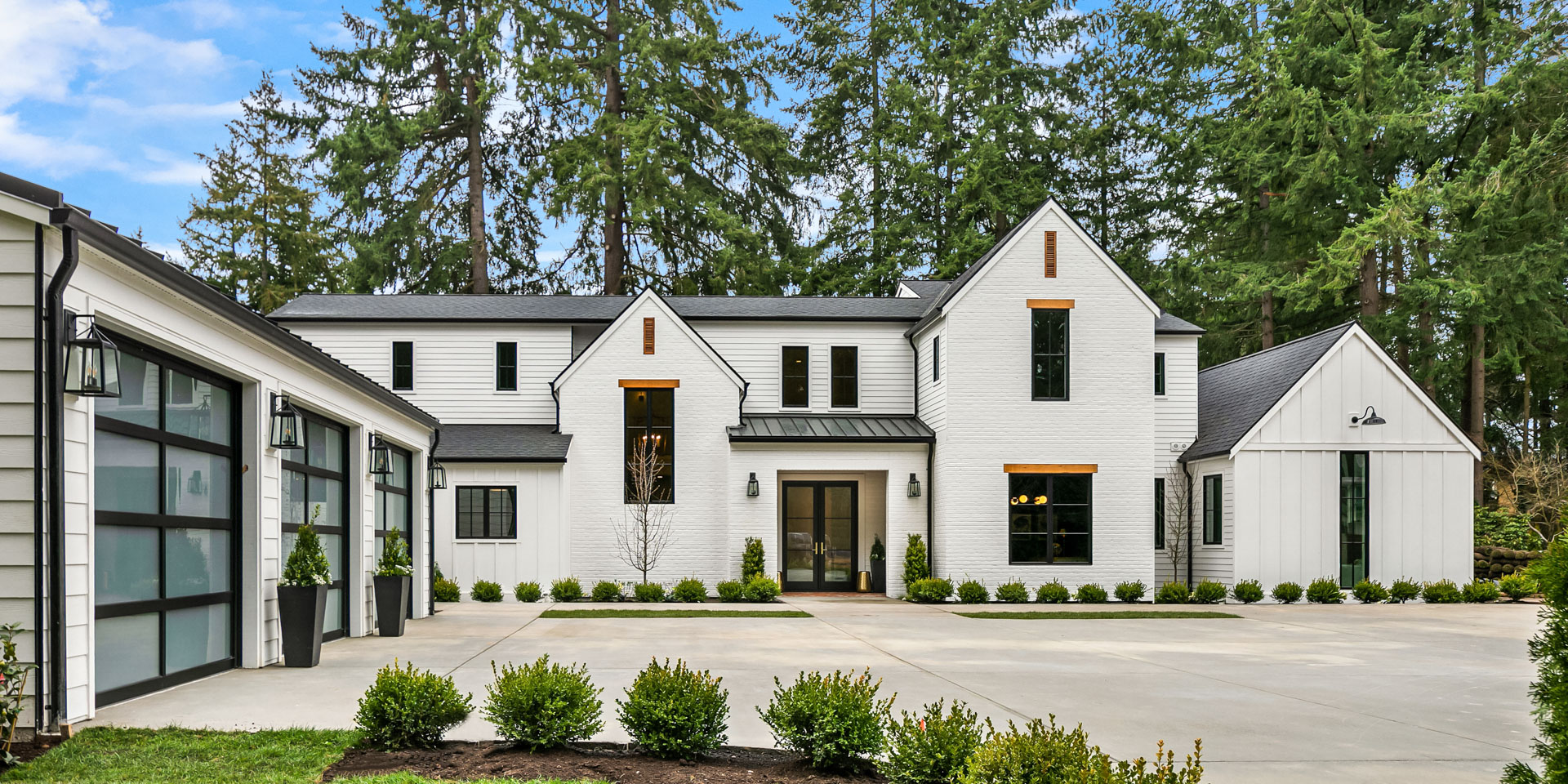 A modern white two-story brick house with black trim, large windows, and a gabled roof. The house is surrounded by tall trees and features a spacious driveway with neatly trimmed bushes lining the front. A three-car garage is attached to the left side of the house.