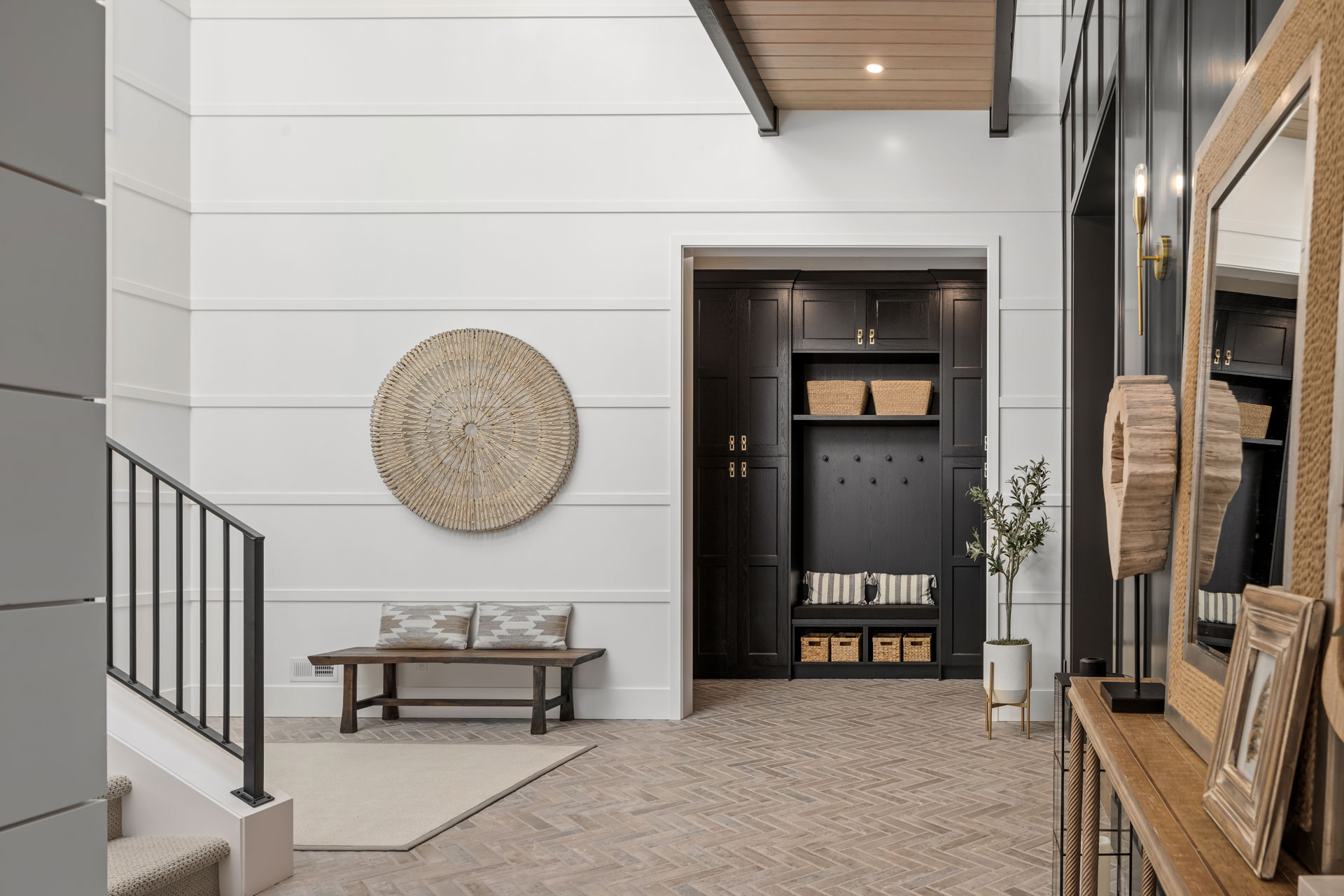 A modern entryway with a high ceiling featuring white shiplap walls and a dark wooden door. A large circular woven wall decor hangs on the left wall above a wooden bench. The floor is herringbone-patterned wood. A small console table holds various decor items.