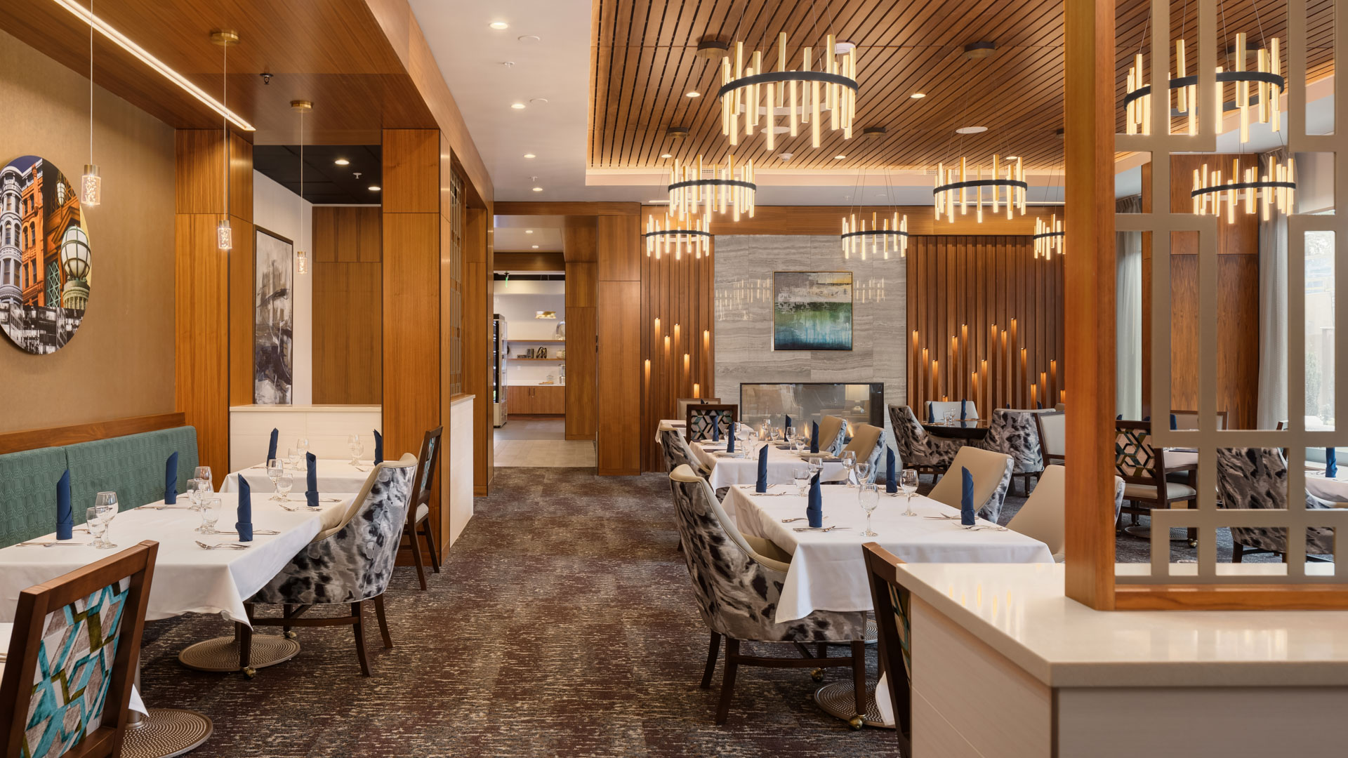 A modern dining room features elegant chandeliers, wooden paneling, and a mix of art on the walls. Tables are set with white linens, blue napkins, and wine glasses. The room includes patterned chairs and a fireplace in the background, creating a warm and upscale ambiance.