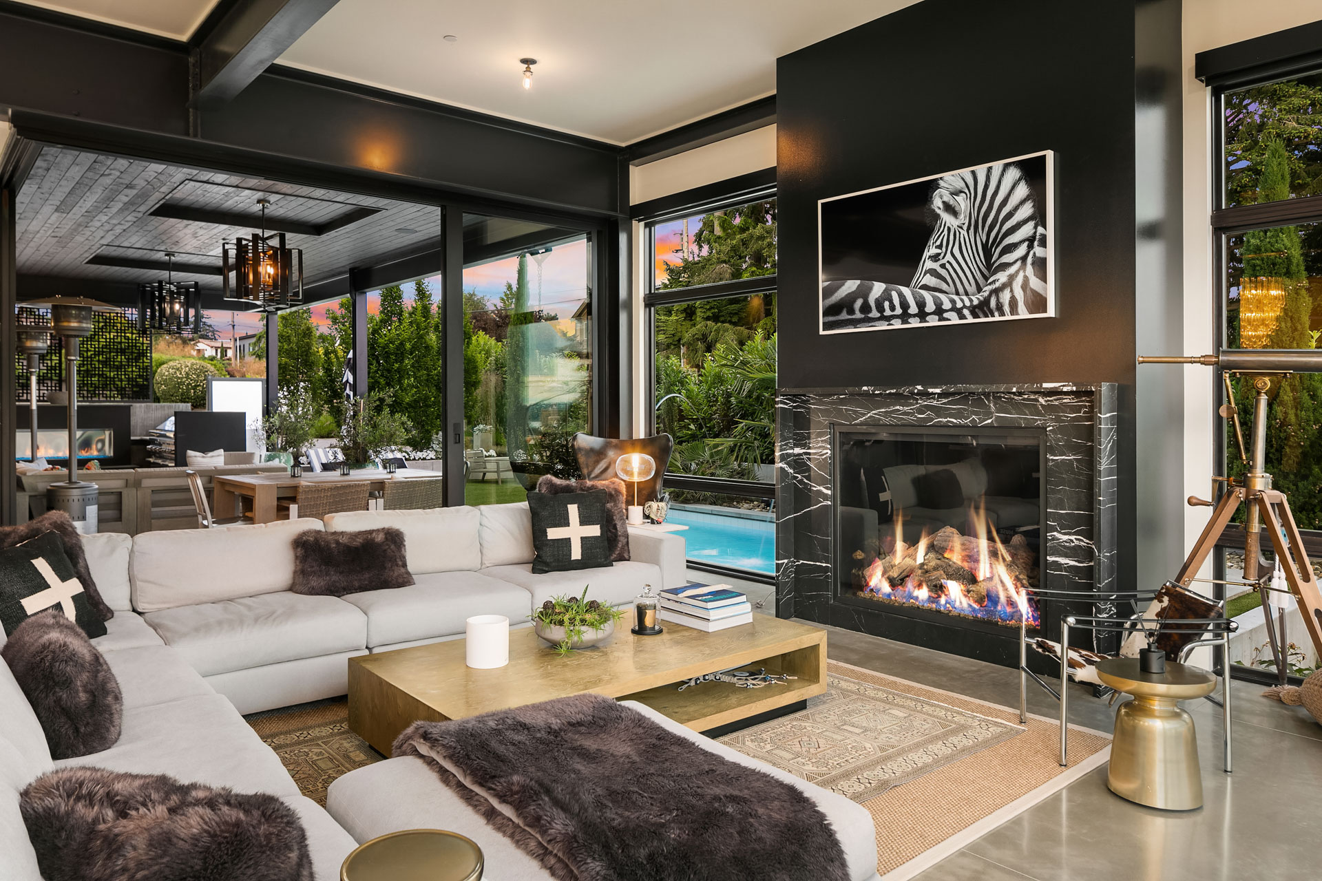 A modern living room featuring a beige sectional sofa with dark throw blankets and pillows around a gold coffee table. There's a lit fireplace with a zebra-themed artwork above it. Large windows overlook a patio with a pool, and a telescope stands nearby.