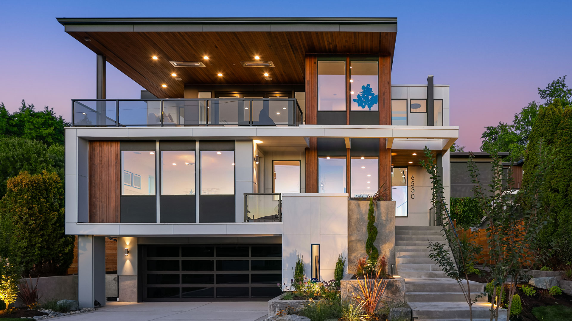 A modern two-story house with a flat roof and large windows, featuring a mix of wood and white exterior accents. It has a spacious balcony, a large garage, and a landscaped front yard with a lit staircase leading to the entrance. The sky is clear and dusk-lit.