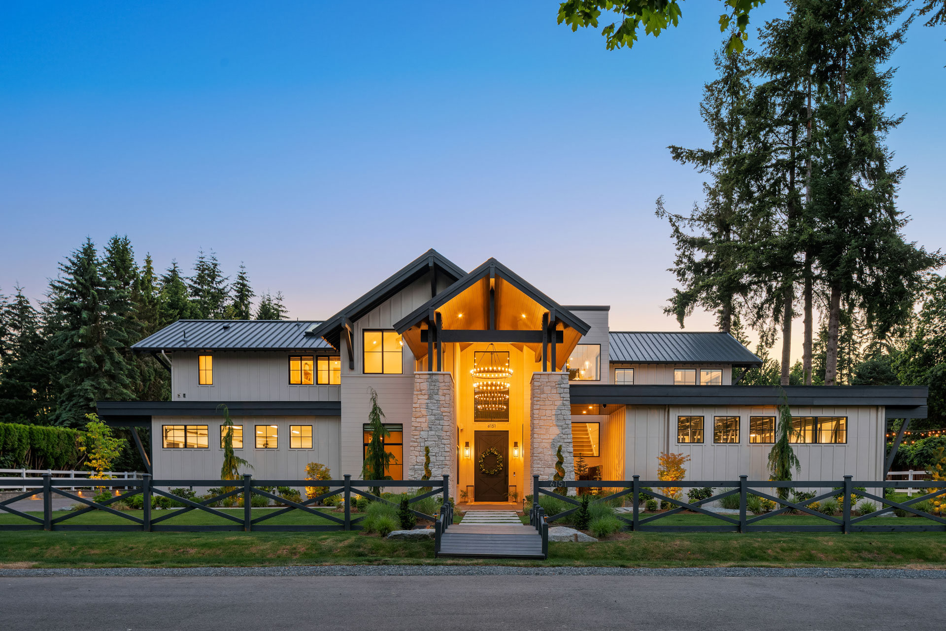 A modern two-story house with large windows and a prominent wooden entrance is framed by lush greenery and tall trees. The house features a mix of stone and wood elements, with warm interior lights glowing through windows at dusk under a clear sky.