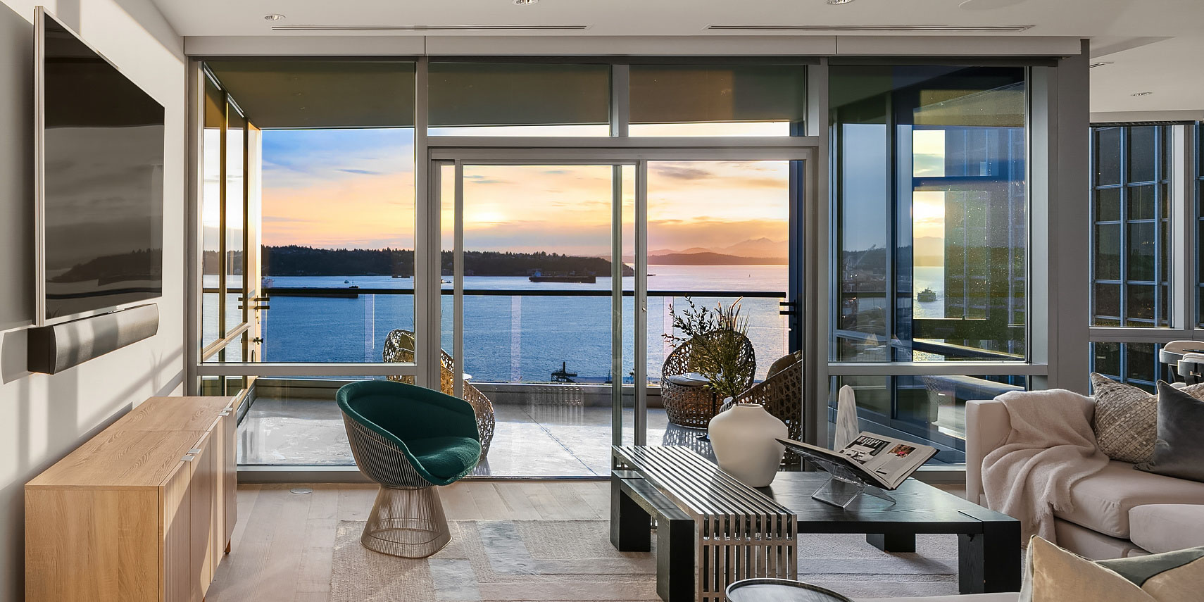 Modern living room with floor-to-ceiling windows overlooking a scenic ocean view at sunset. The room features a TV mounted on the wall, a stylish green chair, a beige sofa, a coffee table, and decorative plants. A balcony with outdoor furniture is visible outside.