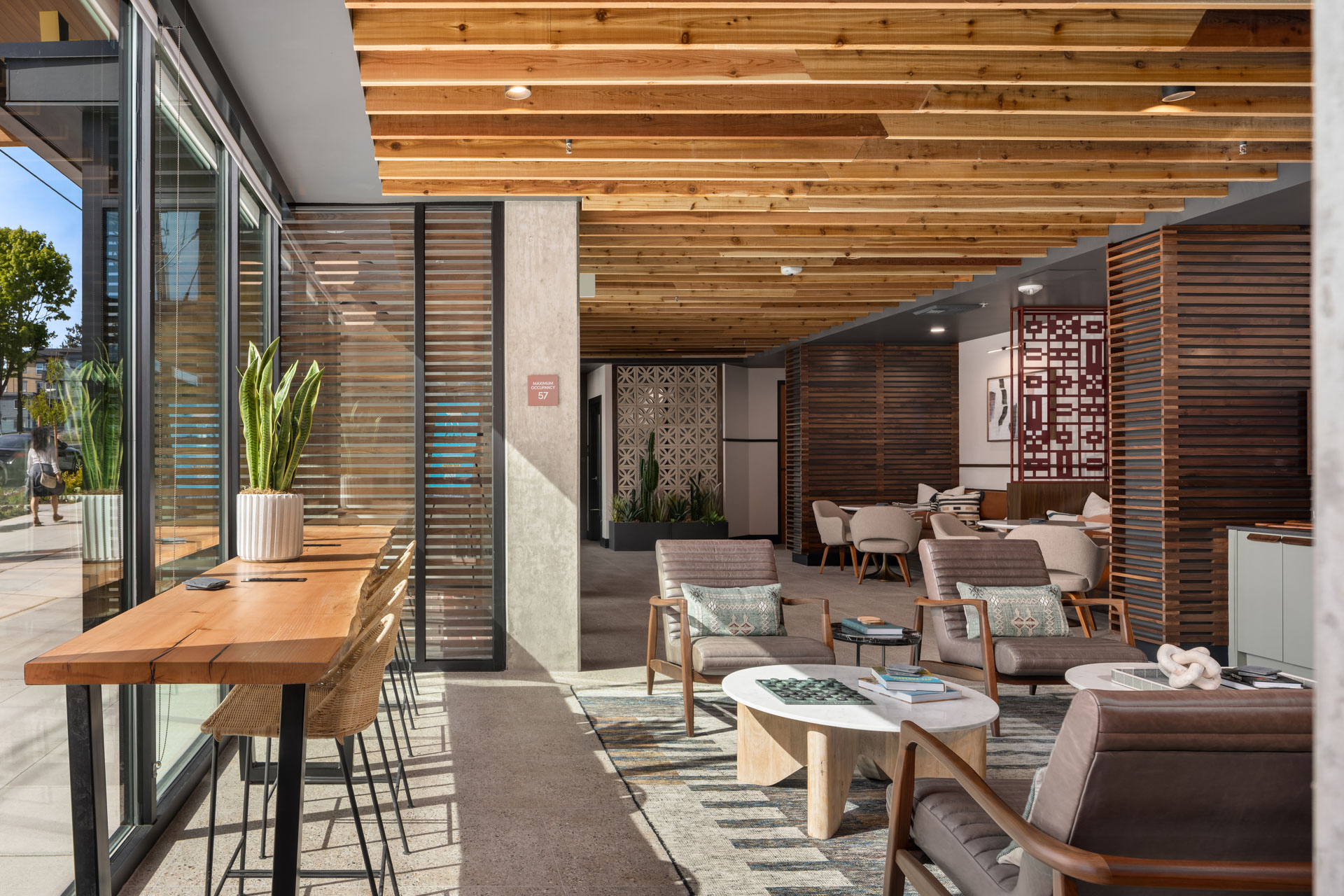 A modern lounge with a sleek, contemporary design featuring wooden ceiling slats and partition walls. The space has several seating areas with wooden chairs, tables, and potted plants by large windows, providing a bright and airy atmosphere.