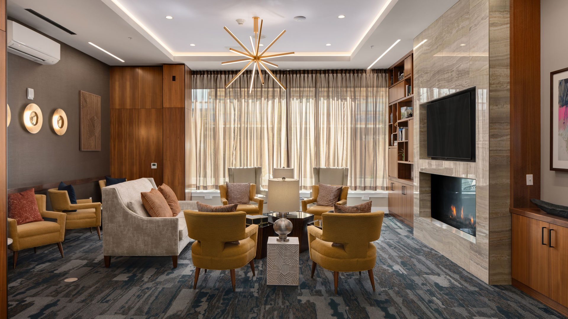 A modern, elegant living room with a central round glass table surrounded by beige and mustard-colored chairs. The room features a fireplace below a mounted TV, wood-paneled walls, large windows with long curtains, and a distinctive starburst chandelier.