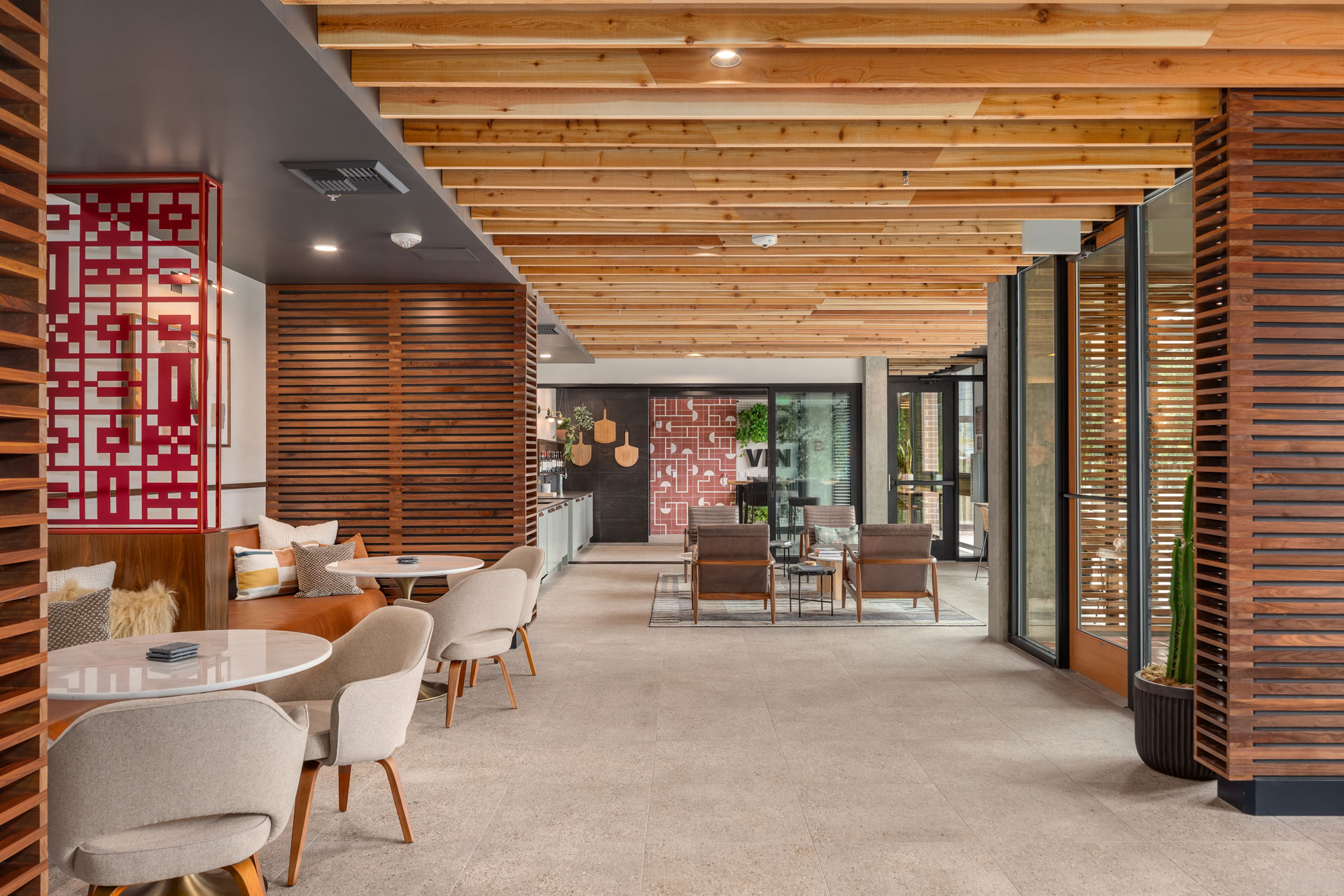 Modern indoor lounge area with wooden slat ceiling and partitions, padded beige chairs around round white tables, and a seating area with leather chairs and a coffee table in the background. Red geometric art decorates the walls, and large windows allow natural light in.