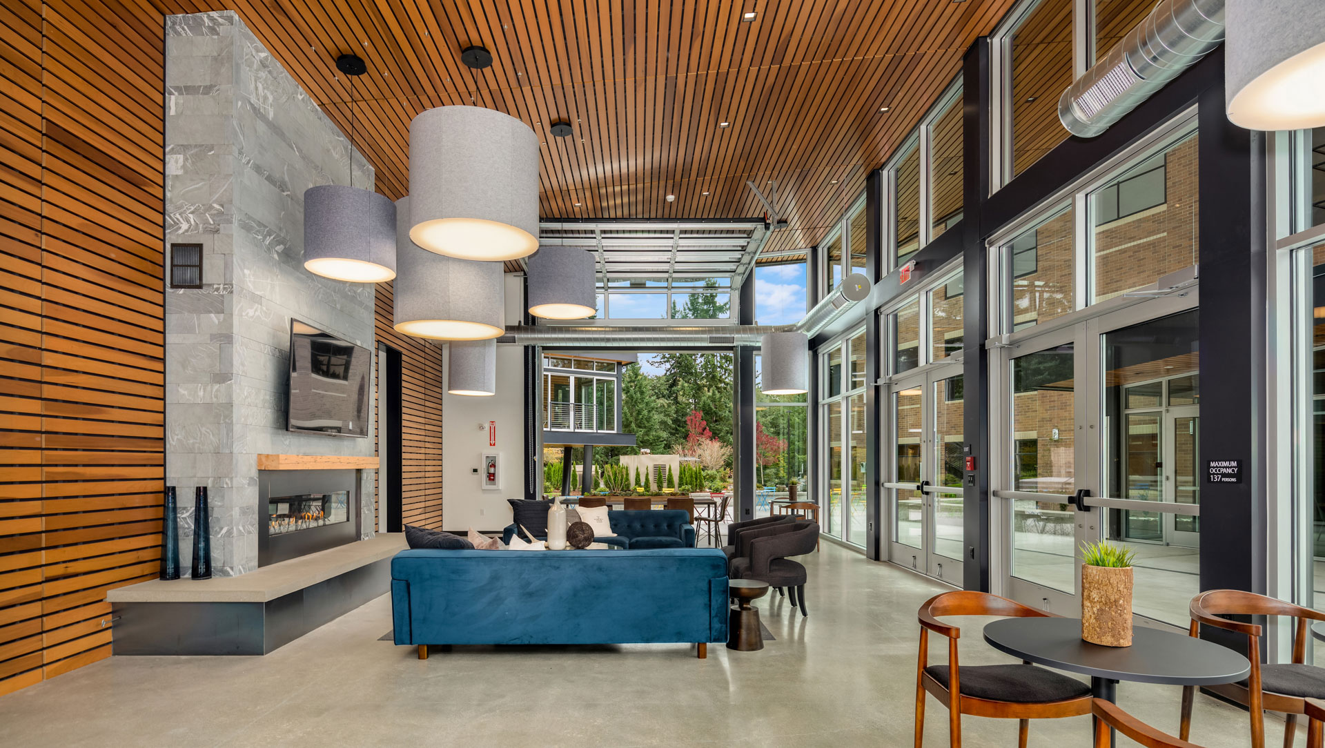 Modern lounge area with wooden ceiling and large windows. The space includes a blue couch, armchairs, a fireplace, and contemporary hanging lights. There is a round table with two wooden chairs in the foreground and a view of greenery and buildings outside.