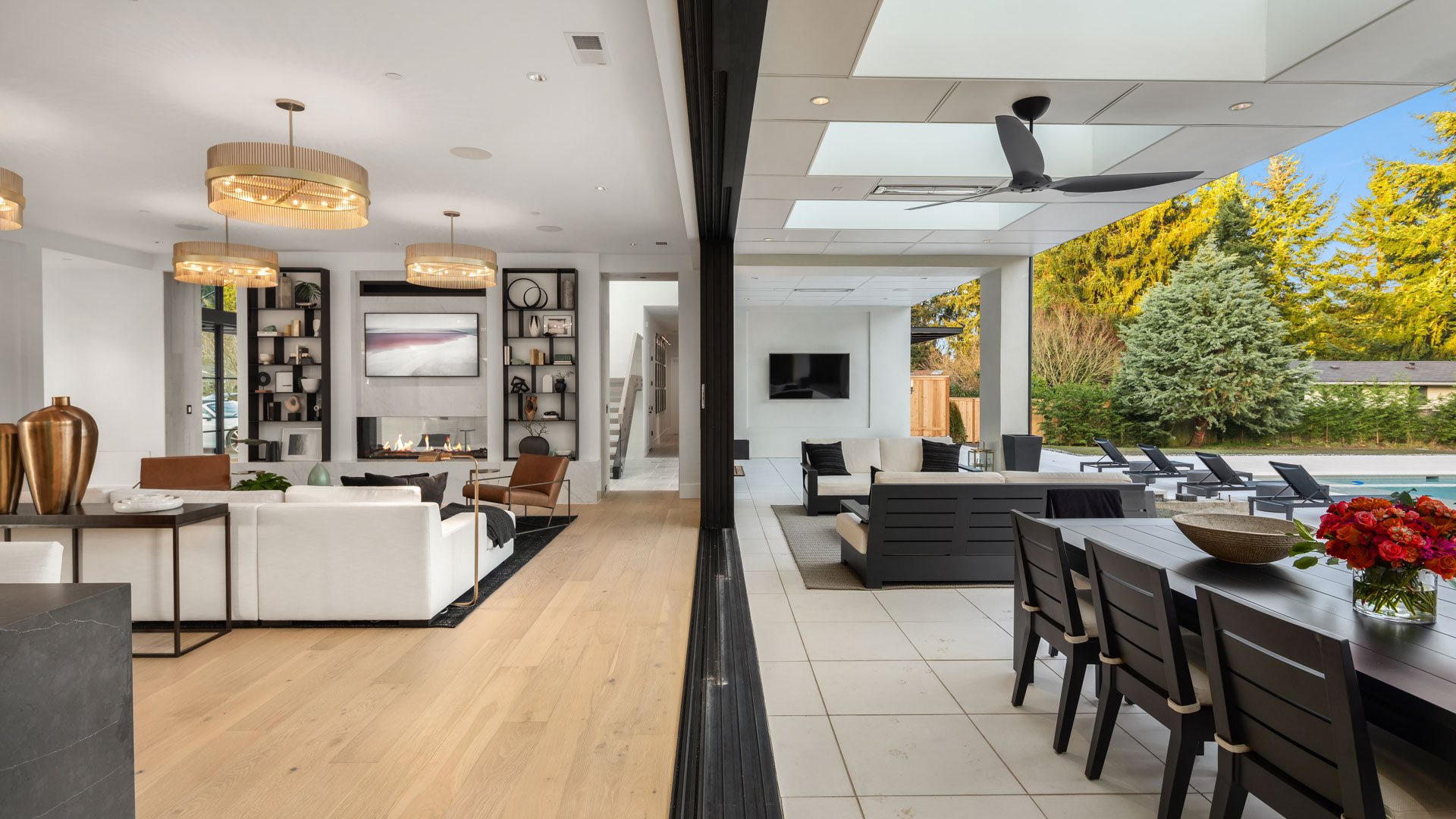 A modern home split view showing a contemporary living room on the left with neutral tones, a fireplace, and stylish decor, and an outdoor patio on the right with a dining table, seating area, ceiling fan, and a swimming pool in the background.