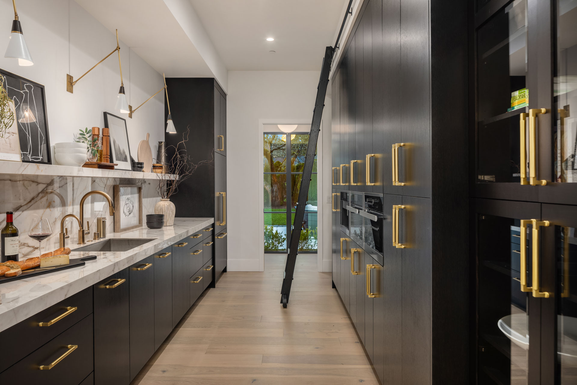 A modern, narrow kitchen with black cabinets, gold handles, and a marble countertop. A sliding ladder is attached for access to higher storage. The kitchen features various plants, decorative items, a wine bottle, and a cheese platter. A door at the end provides outside access.