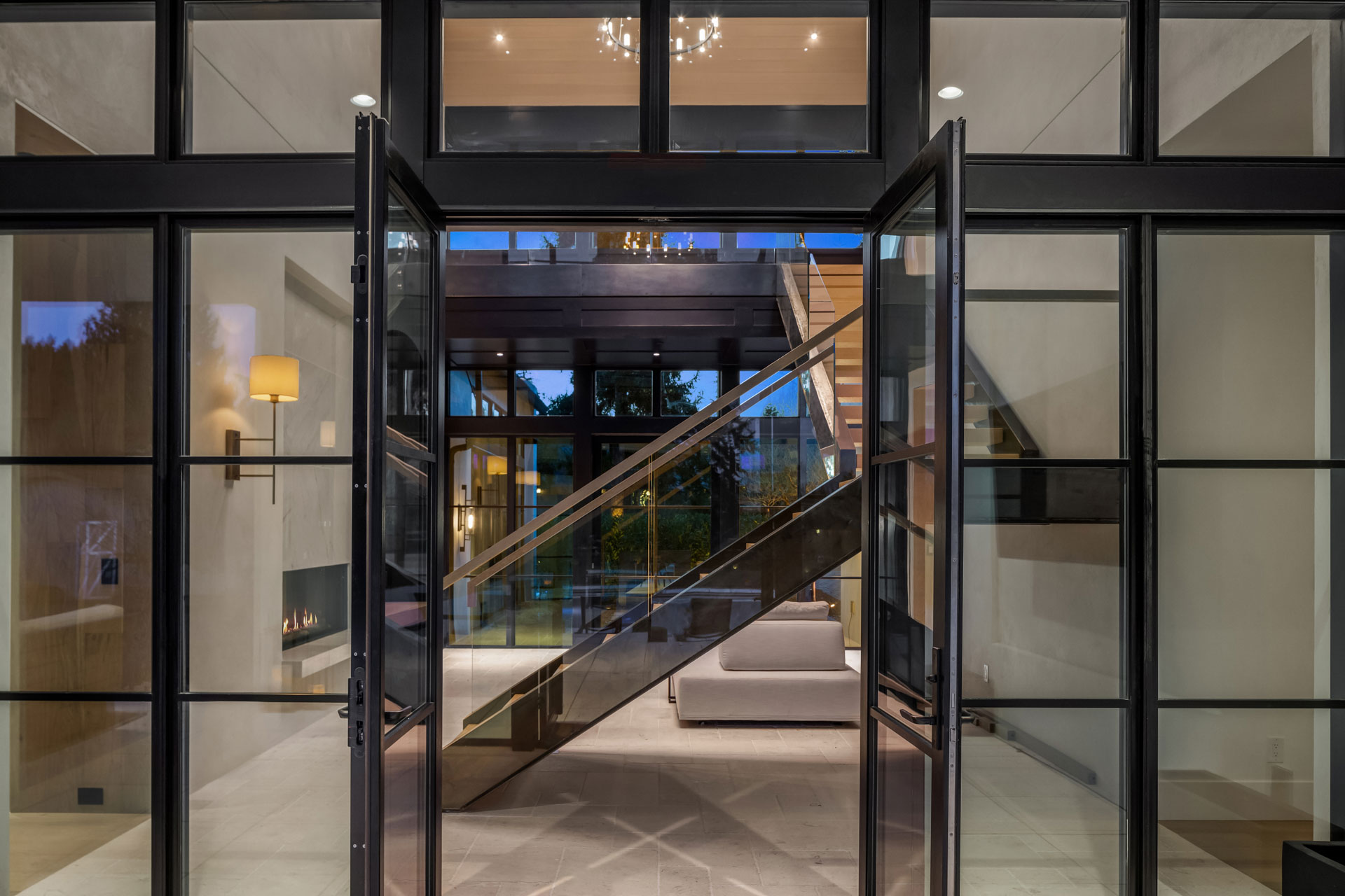 A modern interior entrance features floor-to-ceiling glass doors that open into a spacious living area with sleek, contemporary furniture. A staircase with glass railings ascends to an upper level, and a large chandelier hangs from the ceiling.