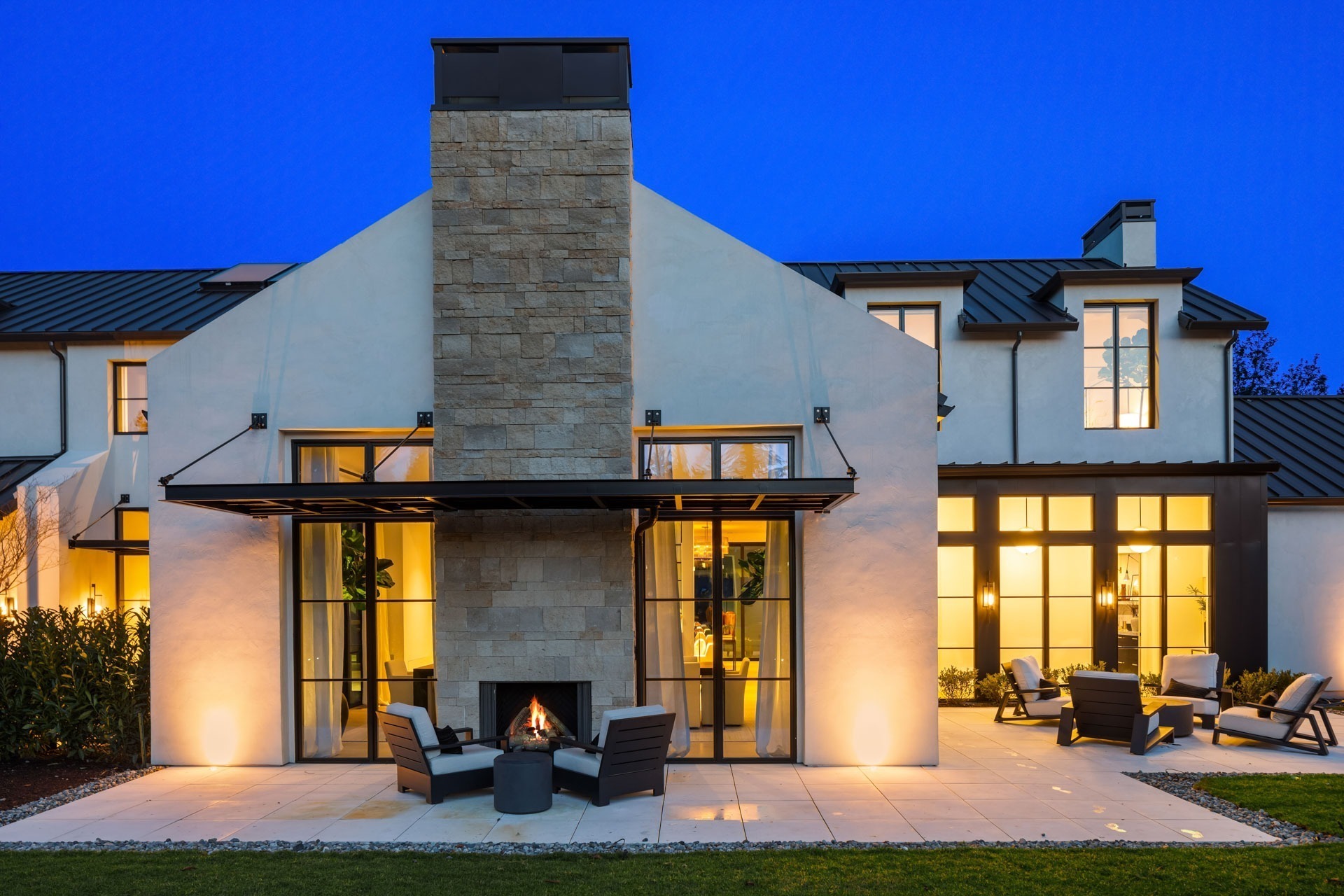 A modern house with a pitched roof and large windows, illuminated at dusk. The exterior features a central chimney with an outdoor fireplace surrounded by patio chairs, set on a spacious tiled patio area. The sky is a clear deep blue.