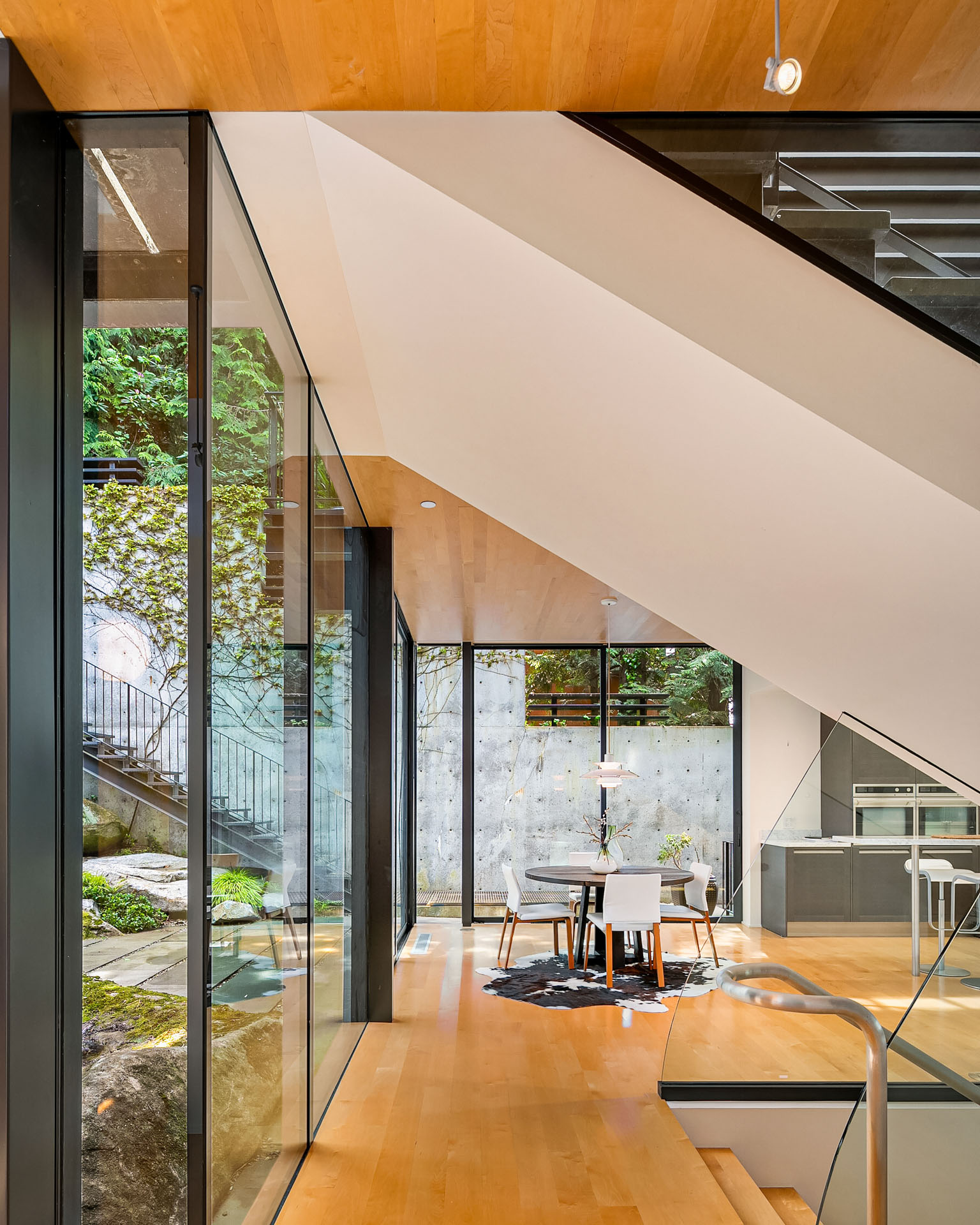 Interior view of a modern home with large floor-to-ceiling windows. A dining area with a rectangular wooden table and white chairs sits beneath a staircase. The space is bright with natural light, showcasing wooden floors and a garden visible through the glass walls.