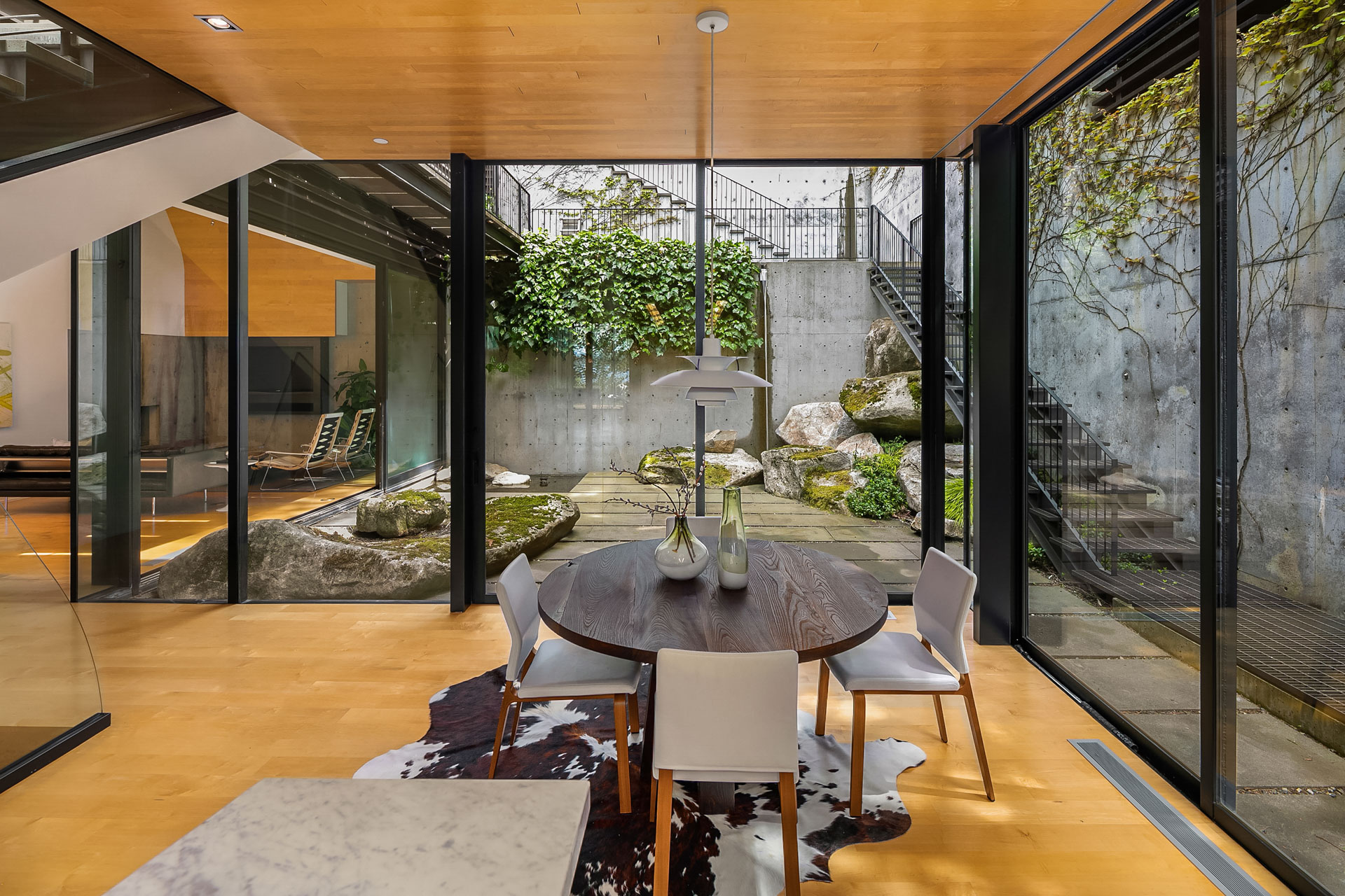 A modern dining room with a wooden ceiling and floor, featuring a round table with white chairs. Large glass doors and windows offer a view of a landscaped courtyard with stone steps and lush greenery. A cowhide rug lies beneath the table, and the space is brightly lit.