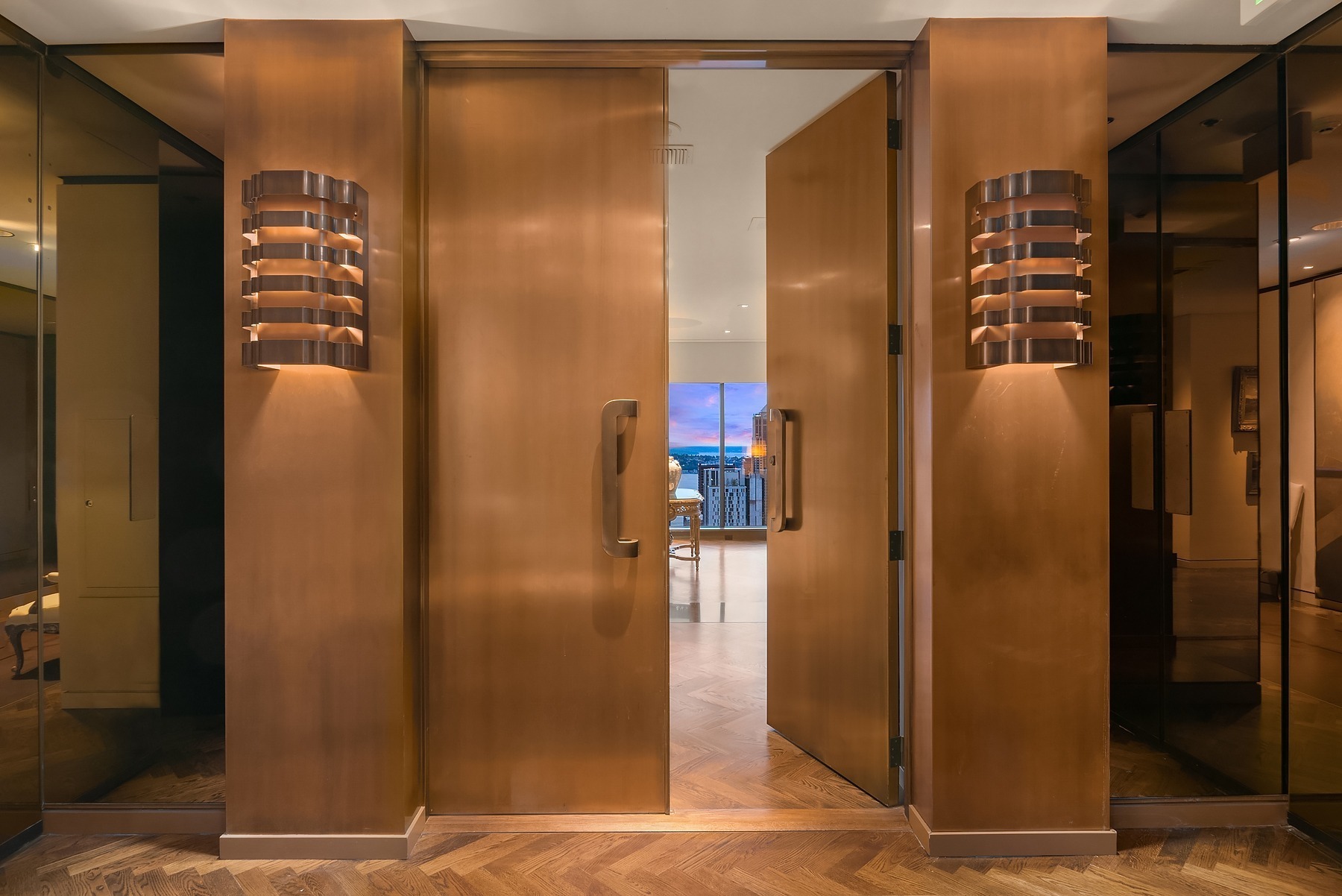 Open wooden double doors with modern wall sconces leading to a room with a view of a sunset. The floor is herringbone-patterned wood, and the walls are mirrored, reflecting the warm lighting. The scene evokes a sense of elegance and warmth.