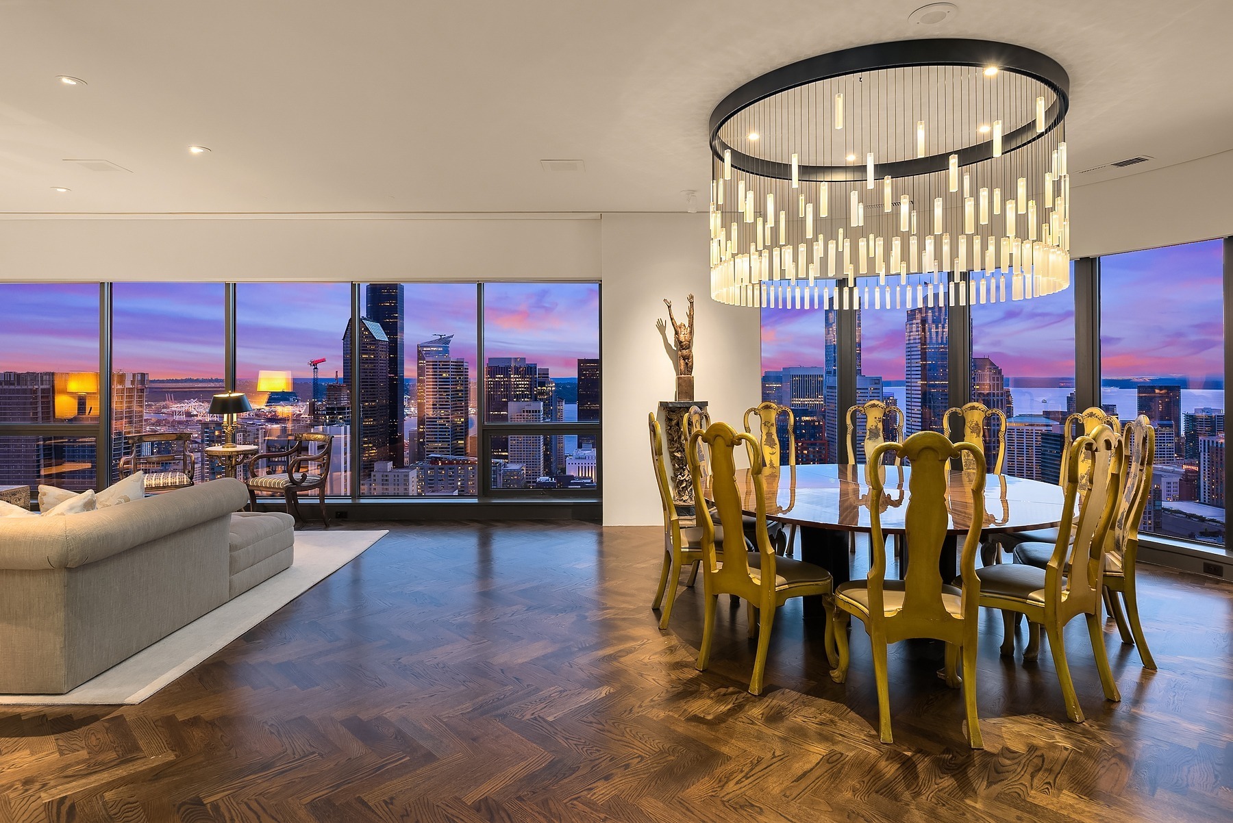 A luxurious living and dining area in a modern high-rise apartment overlooking a city skyline at sunset. The room features a cream sofa, a round dining table with elegant chairs, a large chandelier, and floor-to-ceiling windows showcasing the vibrant cityscape.