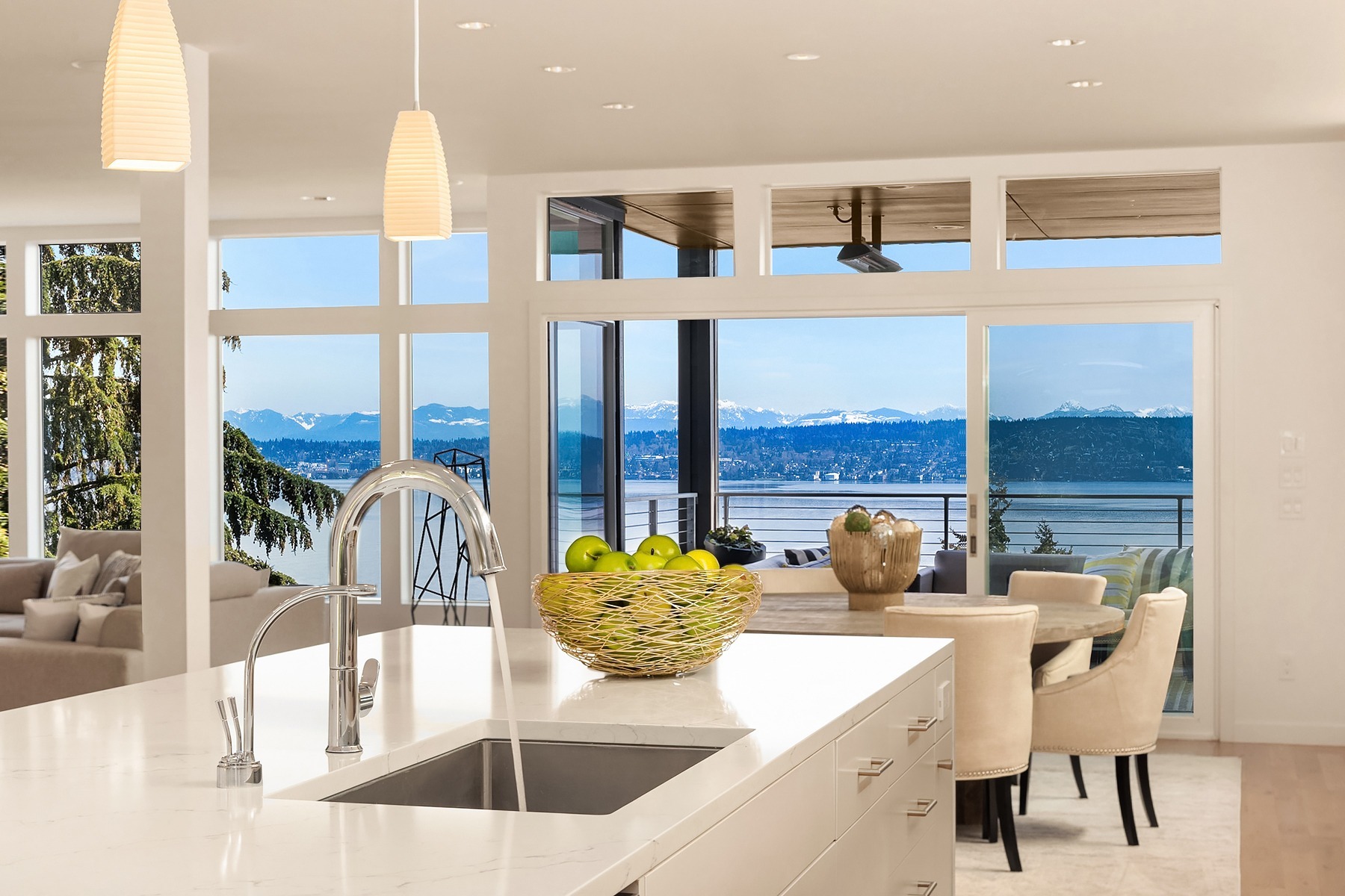 A modern kitchen with a white countertop featuring a sink and a basket of green apples. The kitchen opens into a dining area with a view of a lake and mountains through large windows. Beige chairs and a glass table are in the dining area, with a cozy living room visible to the left.