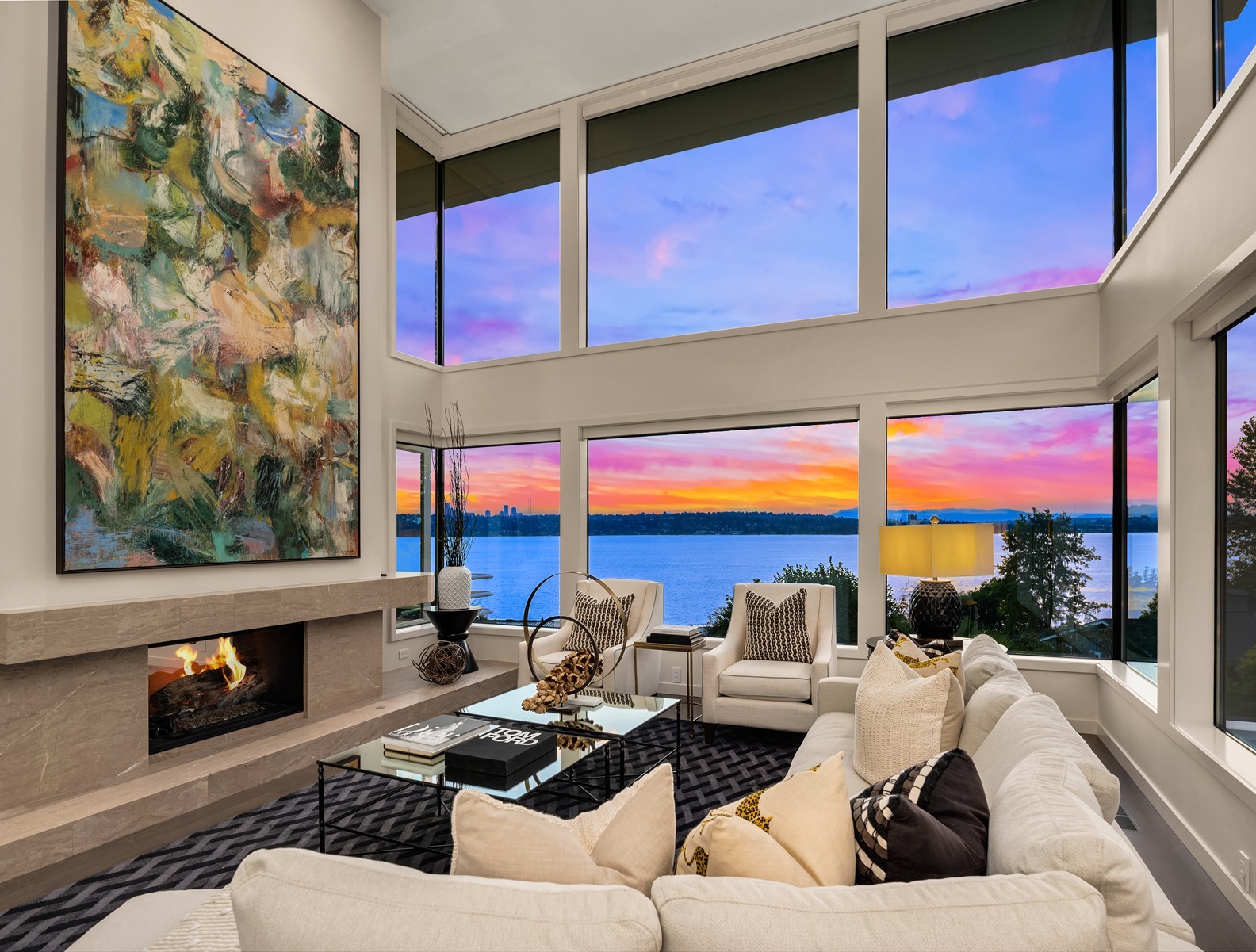 A modern living room features floor-to-ceiling windows showcasing a stunning lake view and vibrant sunset. The room has a large sectional sofa with decorative pillows, a glass coffee table, a fireplace with abstract art above it, and stylish accents throughout.