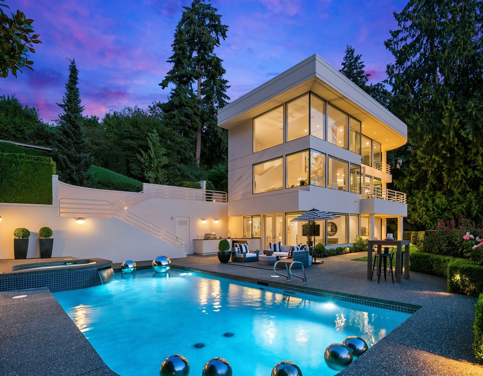 Modern multi-story house with large windows and a stylish patio area illuminated at twilight. The backyard features a swimming pool with metallic water fountains, lounge chairs, an outdoor dining set, and lush greenery, all under a vibrant purple and pink sky.