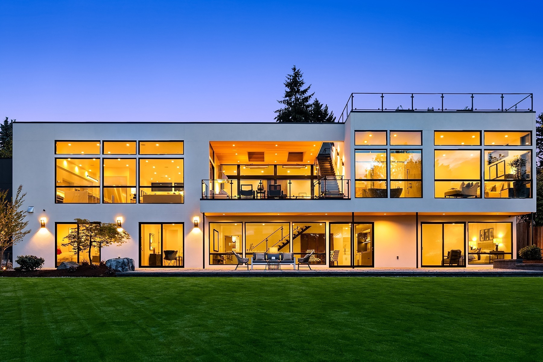 Modern two-story house with extensive glass windows illuminated warmly from the inside, showcasing a minimalist architectural design. The lawn in the foreground is well-maintained, and there's a darkening blue sky in the background.