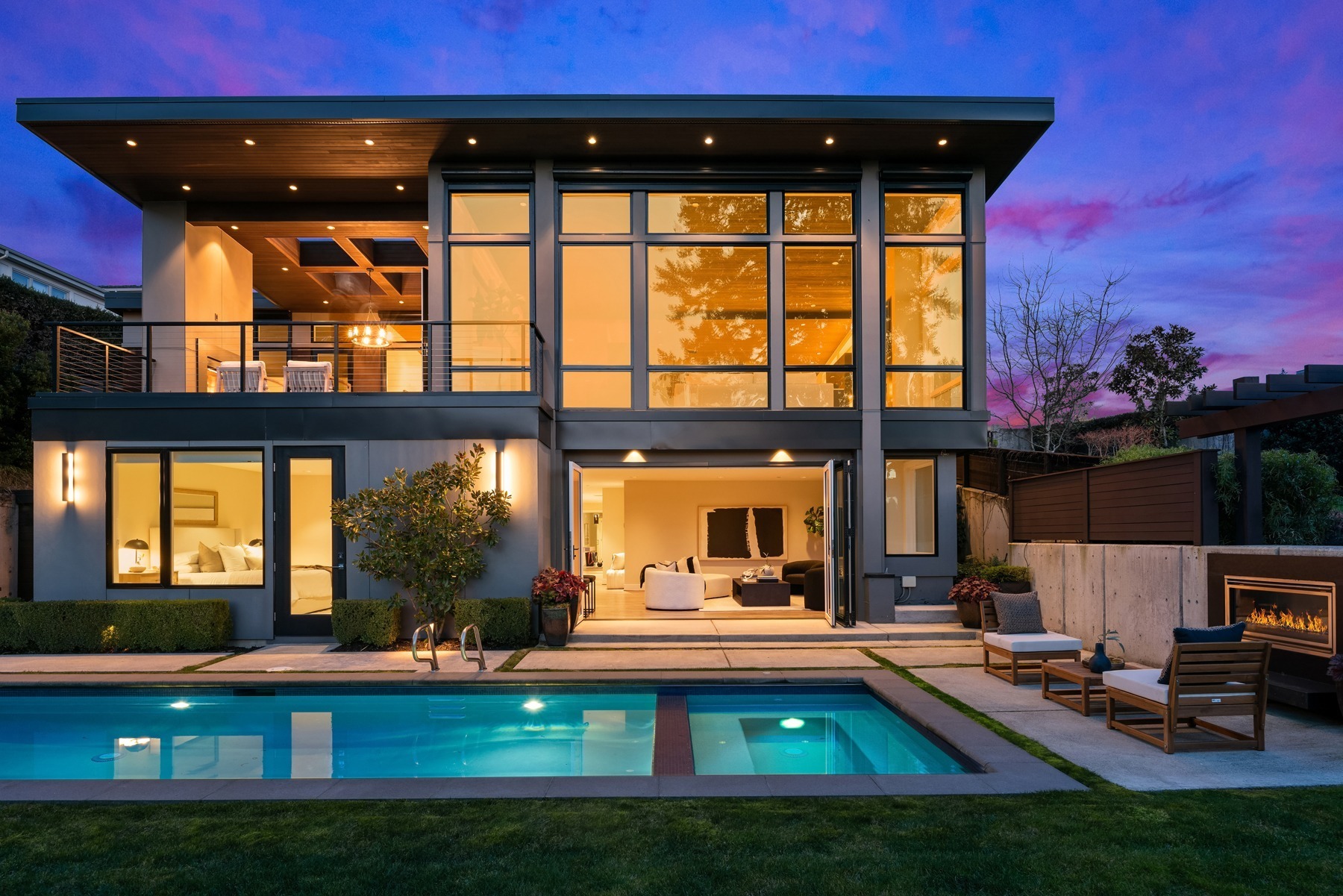 Modern two-story house with large windows, illuminated by soft interior lights at dusk. A pool is in the foreground, with outdoor seating and a fireplace to the right. The sky is a vibrant mix of purple and pink hues, adding to the tranquil ambiance.