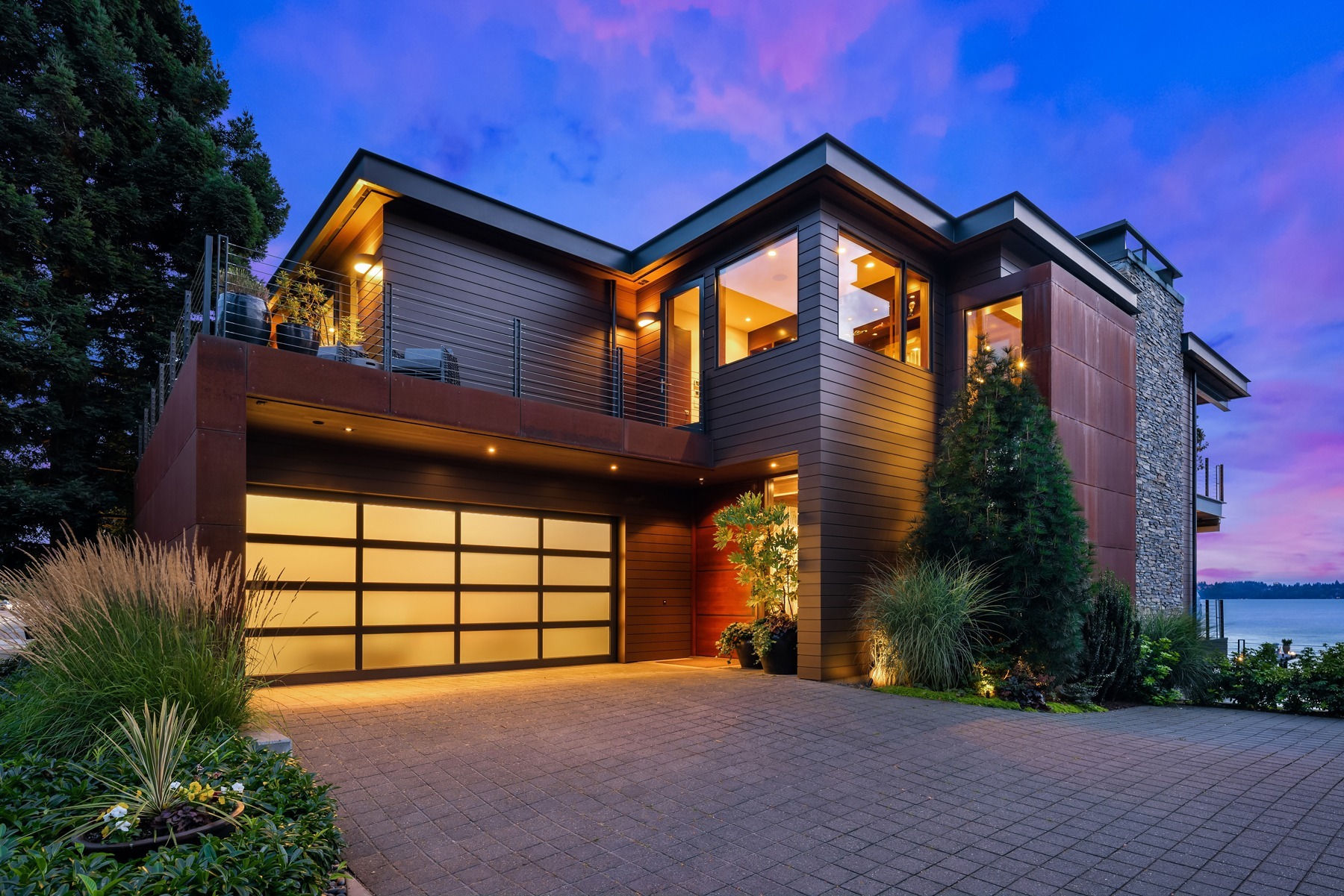 A modern two-story house with large windows, wooden paneling, and a glass-paneled garage door is illuminated by exterior lights during sunset. The well-manicured front yard features various plants and shrubs, and the lake is visible in the background.