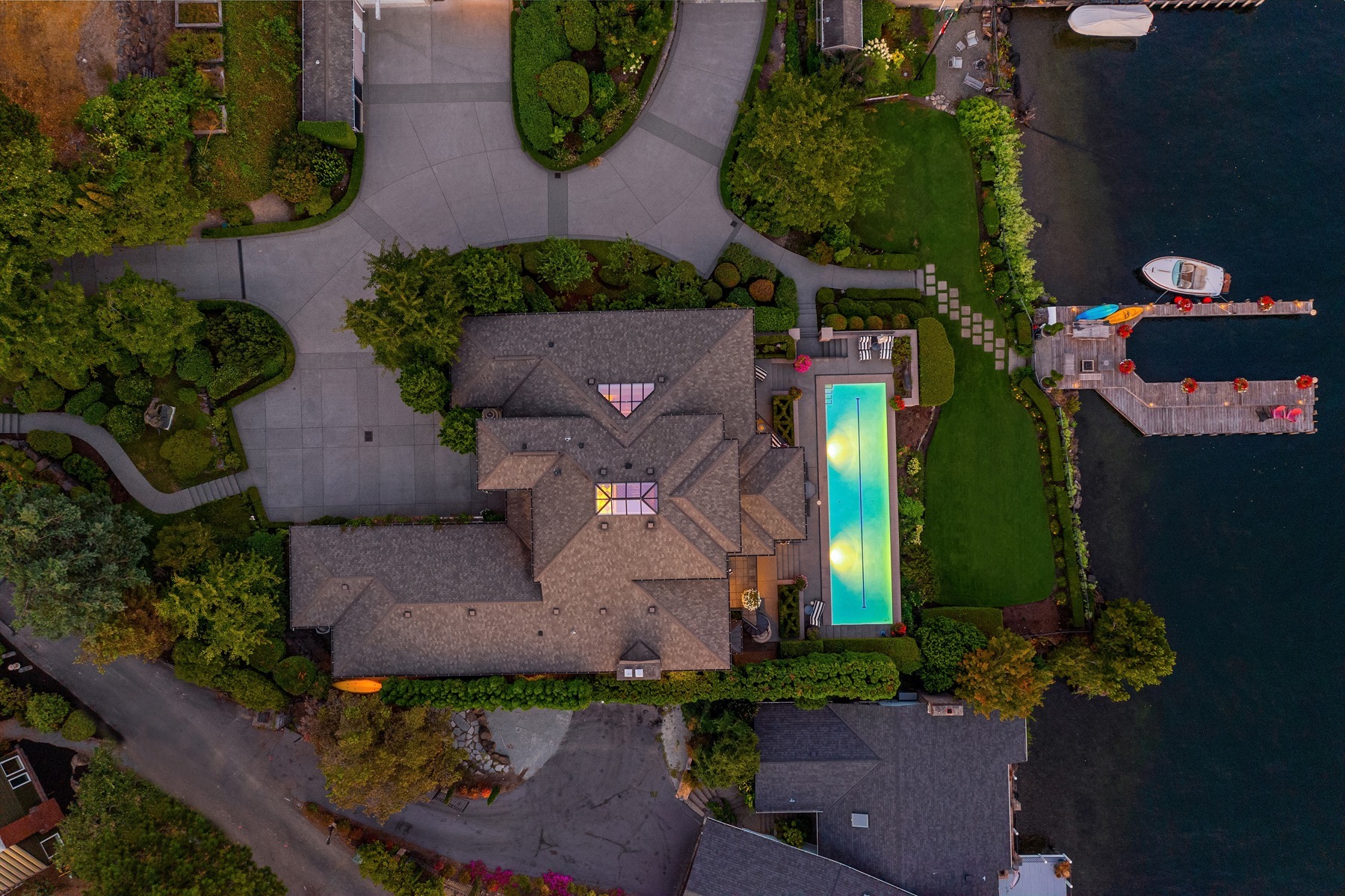 Aerial view of a large house with a complex roof and well-maintained landscaping. The property features a swimming pool with a partially transparent cover, a driveway winding around the house, and a dock leading to a calm body of water with two boats moored.