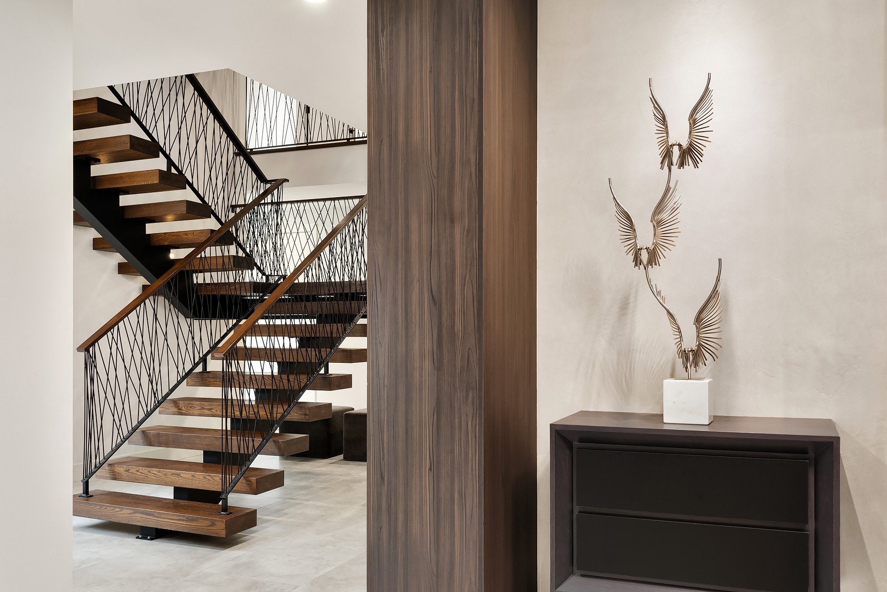 A modern staircase with wooden steps and black railings is seen in an entryway. A beige wall features three decorative metal wing sculptures mounted on it. In the foreground, there is a dark wooden cabinet. The floor is covered in light gray tiles.