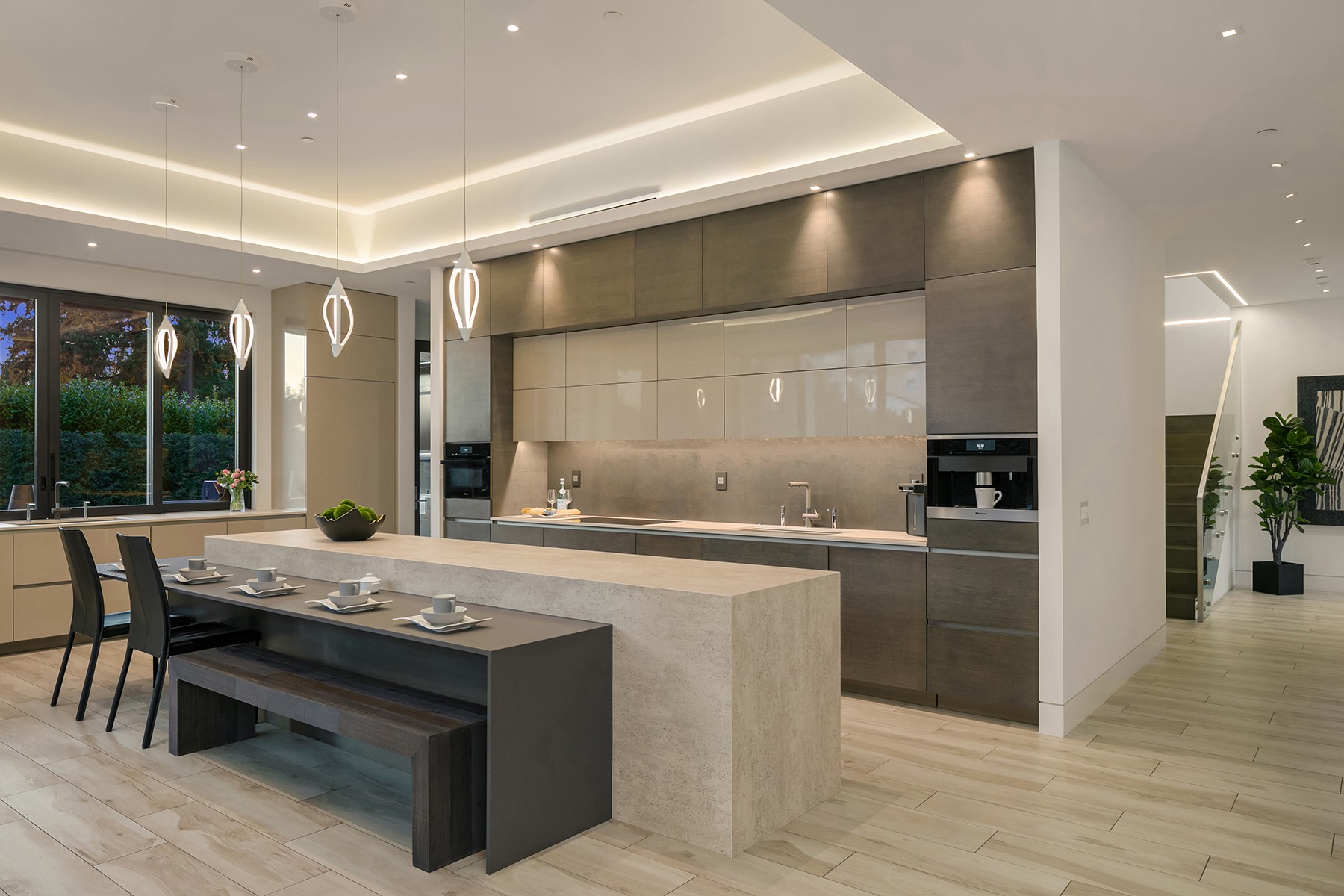 A modern kitchen with minimalist design, featuring sleek cabinets, built-in appliances, a large island with a light countertop, and a dining area with a bench and chairs. Pendant lights hang above the island, and a large window offers a view of a garden.