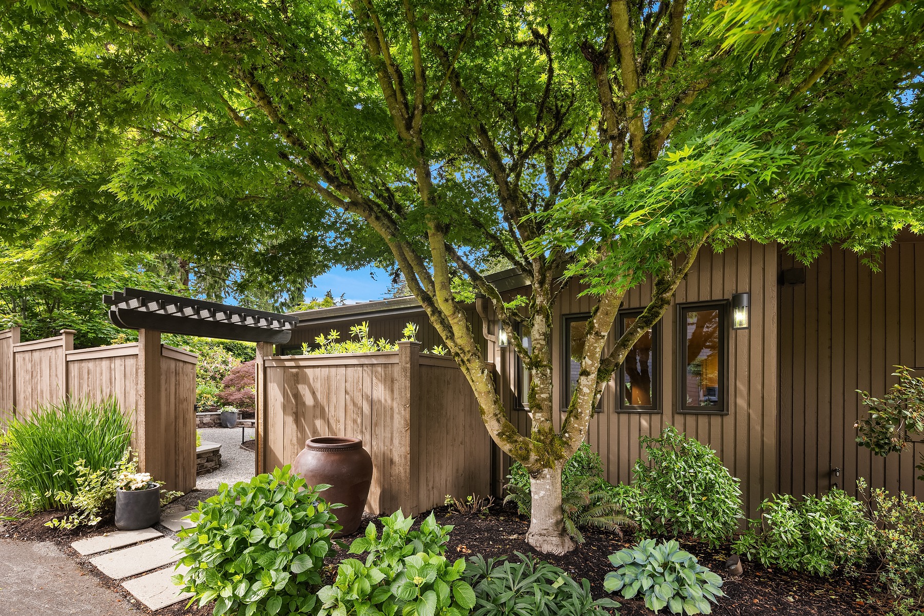 Exterior view of a home featuring wooden siding, large windows, and an inviting garden. A prominent tree with lush green leaves stands in front, adjacent to a large ceramic pot and paved walkway leading to an entry gate. The surrounding garden is well-maintained with diverse plants.