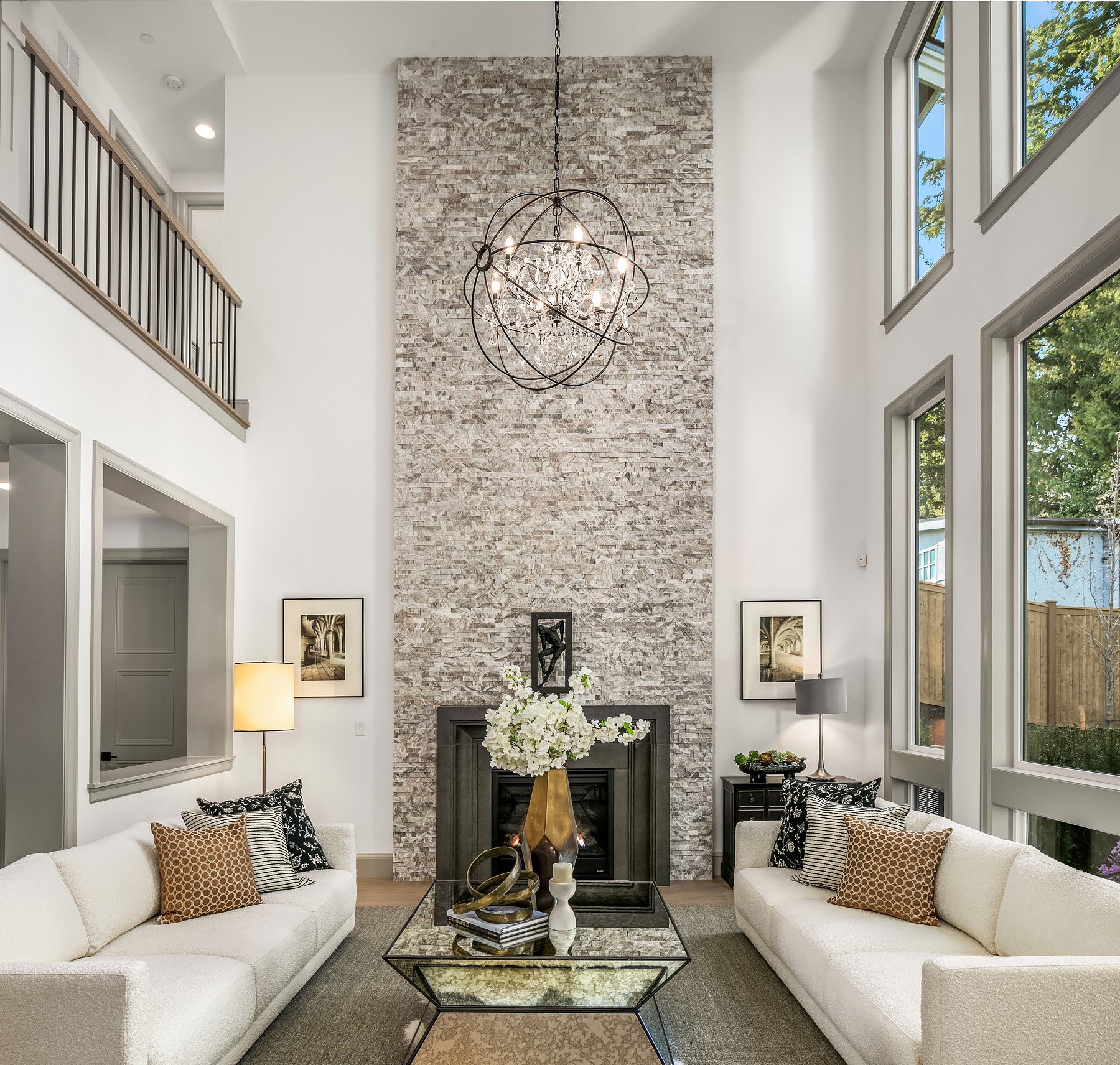 A modern living room with two white sofas facing each other and a glass coffee table in between. A tall stone fireplace extends to the ceiling, adorned with a chic chandelier. Large windows on both sides allow natural light to flood the space.