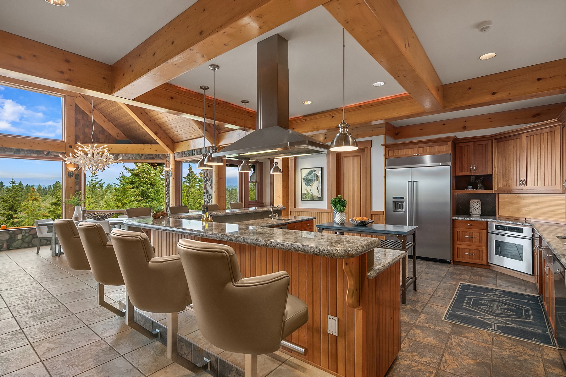 A modern kitchen features a large central island with granite countertops, four beige barstools, stainless steel appliances, and wooden cabinetry. A large window provides a scenic view of greenery and blue skies, and a unique chandelier hangs near the window.