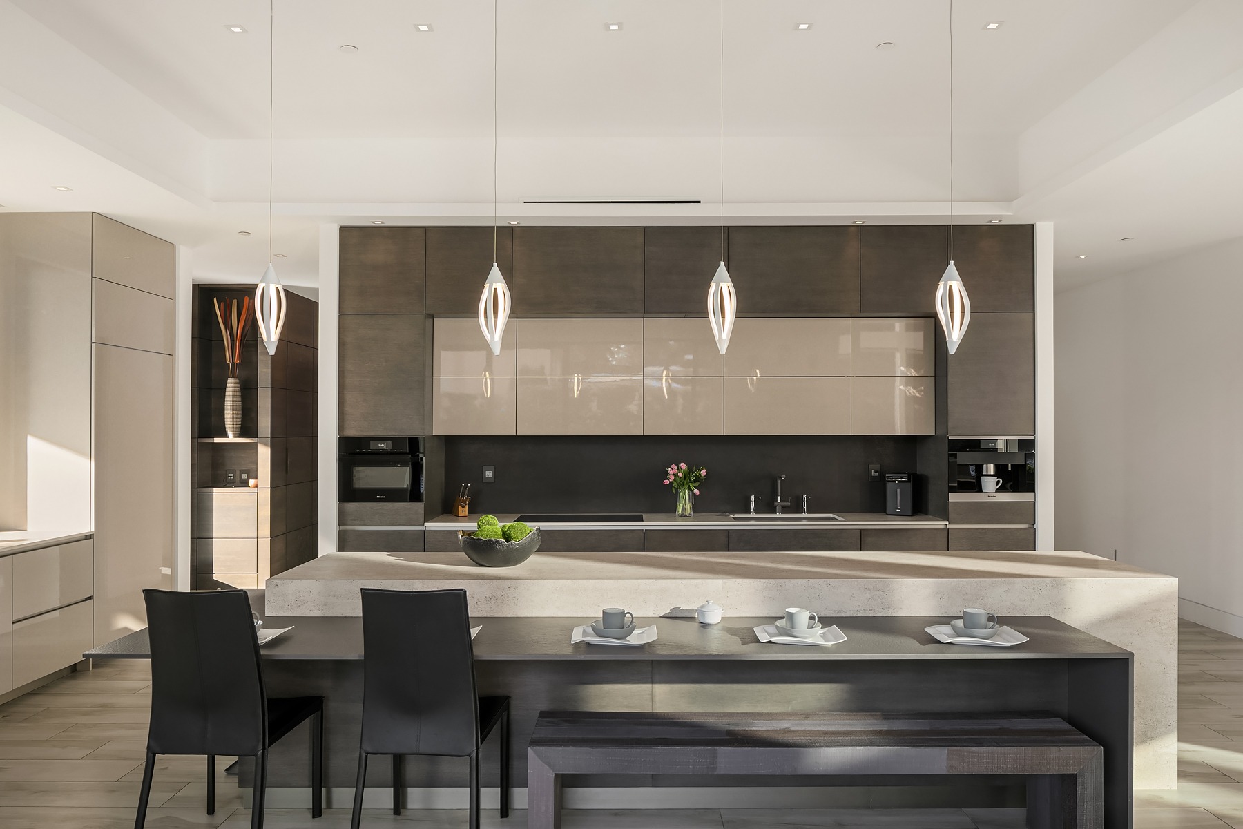 A modern kitchen with sleek cabinetry and a large island featuring four unique pendant lights overhead. The island has a built-in bar with three black chairs and a tray with cups and bowls. The kitchen is decorated with a vase of flowers and a bowl of green apples.