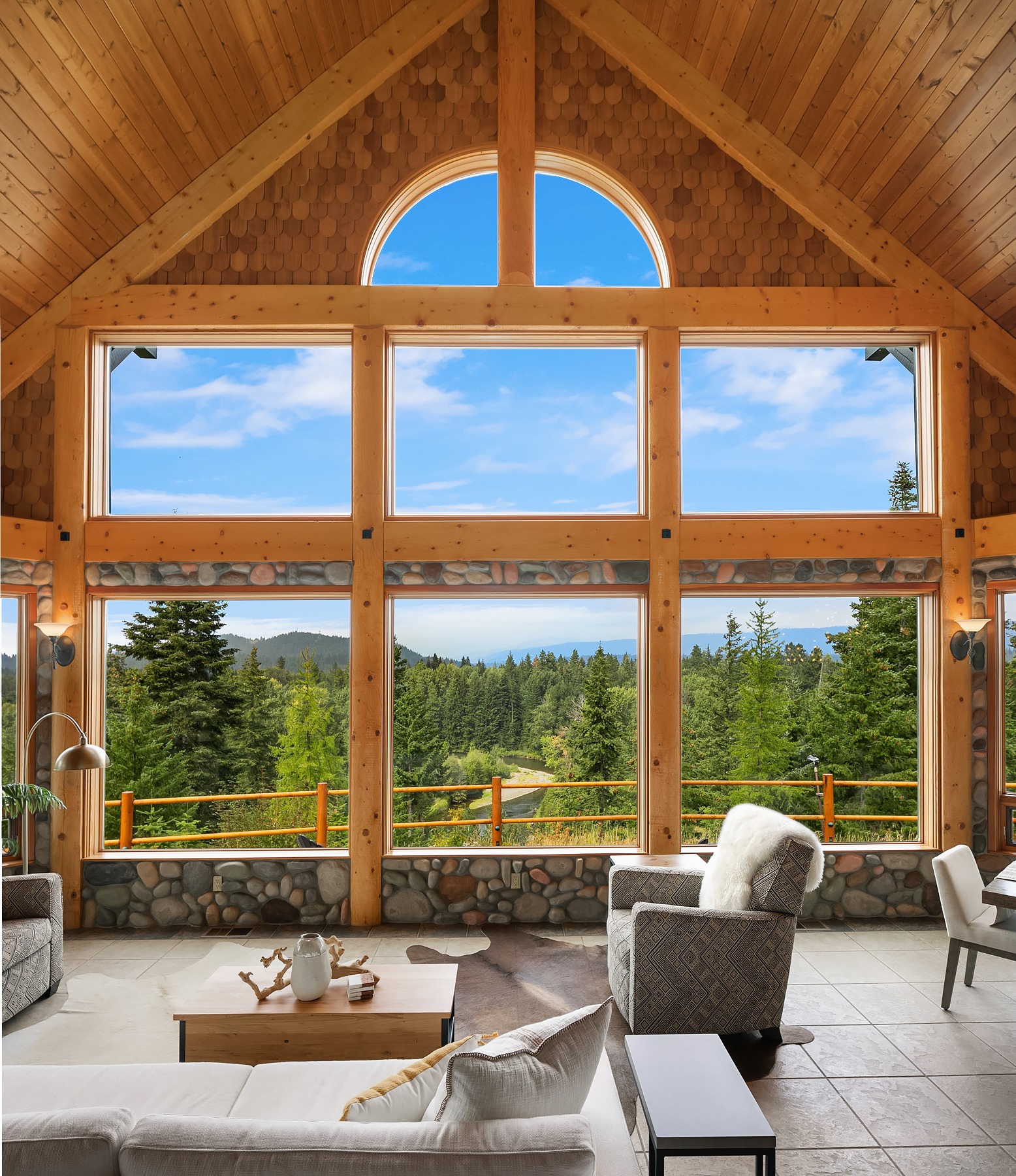 A spacious living room with large floor-to-ceiling windows overlooking a scenic forested landscape. The room features wooden walls and ceiling, a stone wall base, modern furniture, and a high, arch-shaped window. The sky outside is bright blue with scattered clouds.