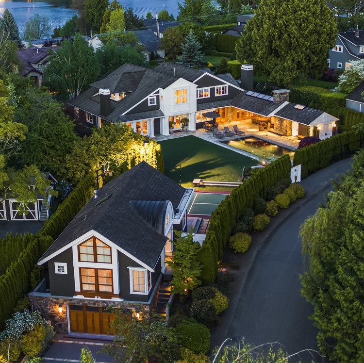 Aerial view of a luxurious property featuring two large houses with dark roofs and white trim, surrounded by dense greenery and landscaped gardens. The compound includes a pool area and overlooks a serene body of water in the background.